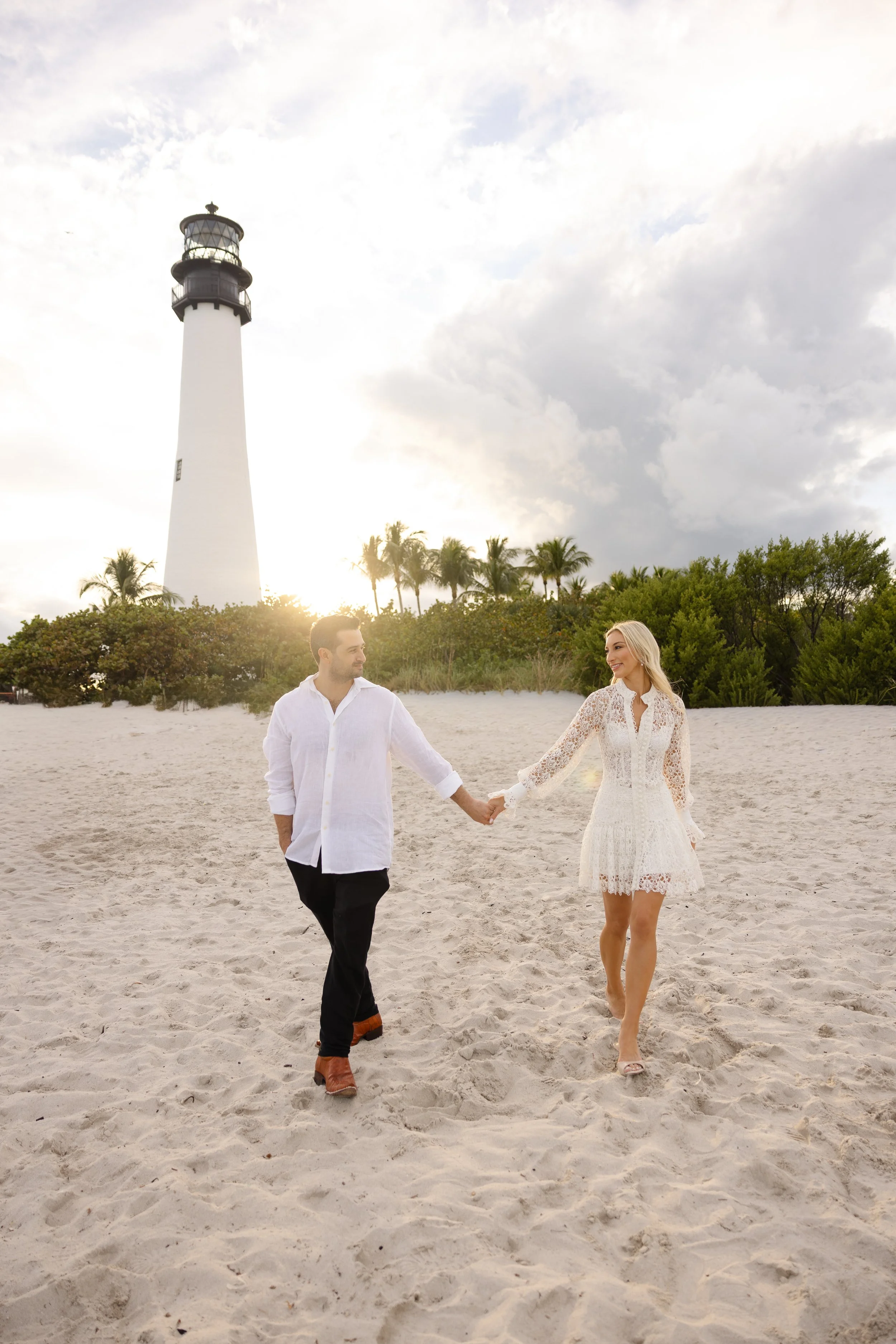 A couple holding hands and walking on a sandy beach with a lighthouse, palm trees, and green bushes in the background, during sunset or sunrise.