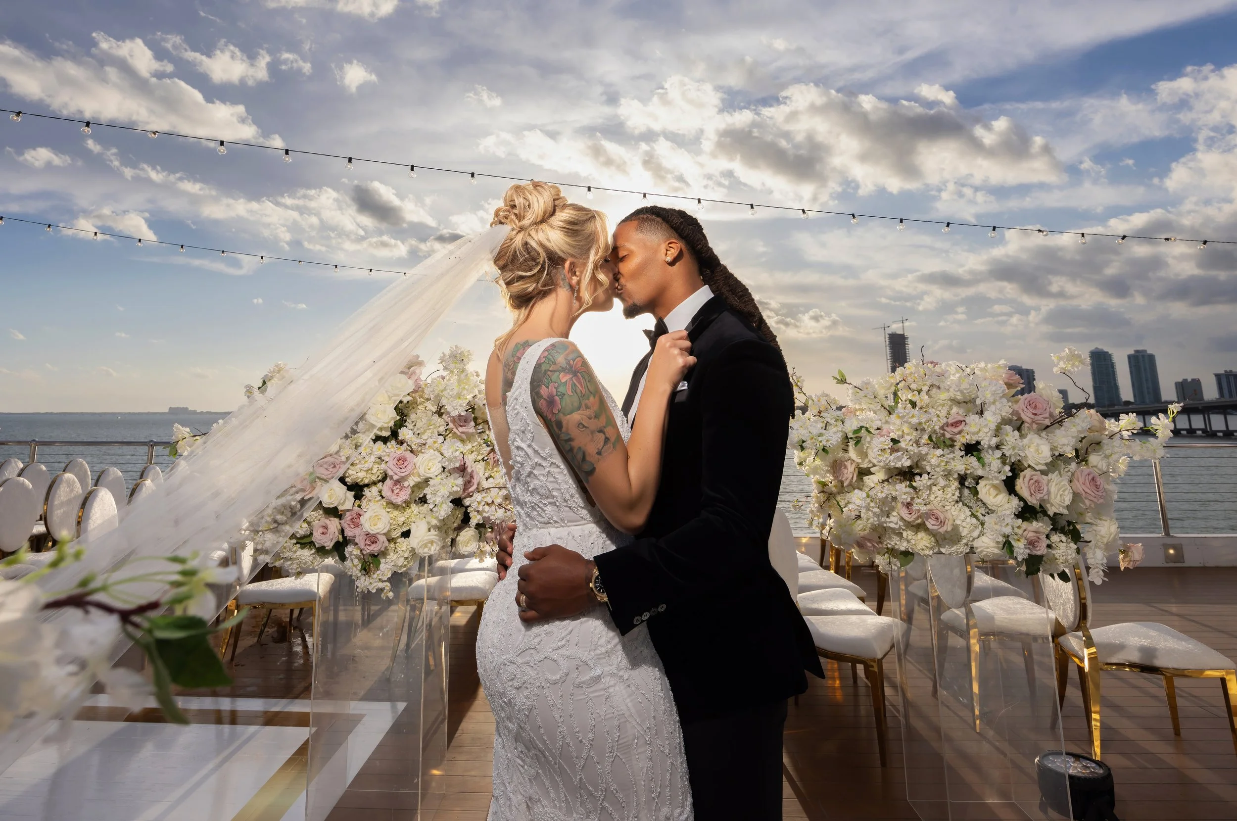 A couple getting married on an outdoor dock, kissing, surrounded by floral arrangements with a city skyline and water in the background.