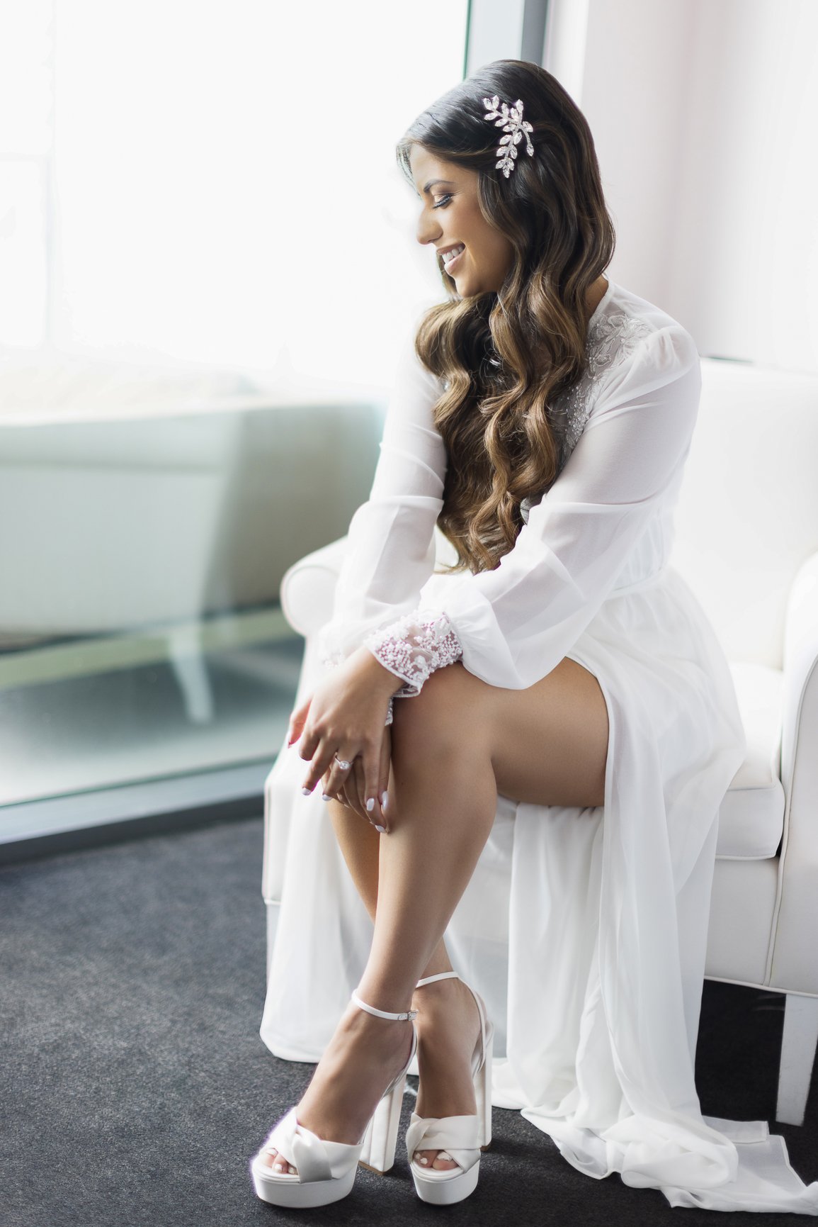 A woman with long wavy hair, wearing a white robe with lace details and white high heels, sitting on a white chair in a bright room.
