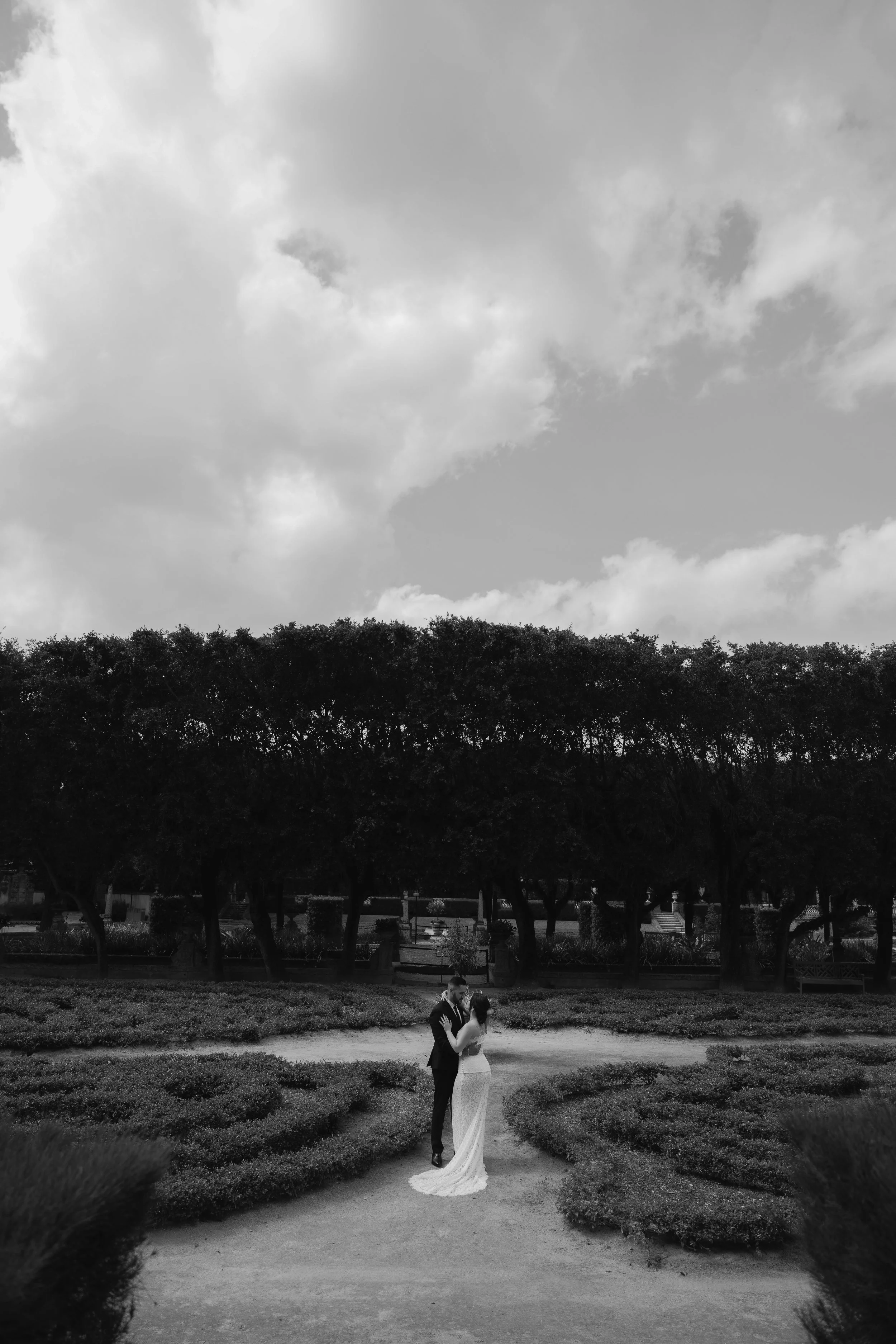 A couple in wedding attire sharing a kiss in a garden with trees in the background and a cloudy sky above.