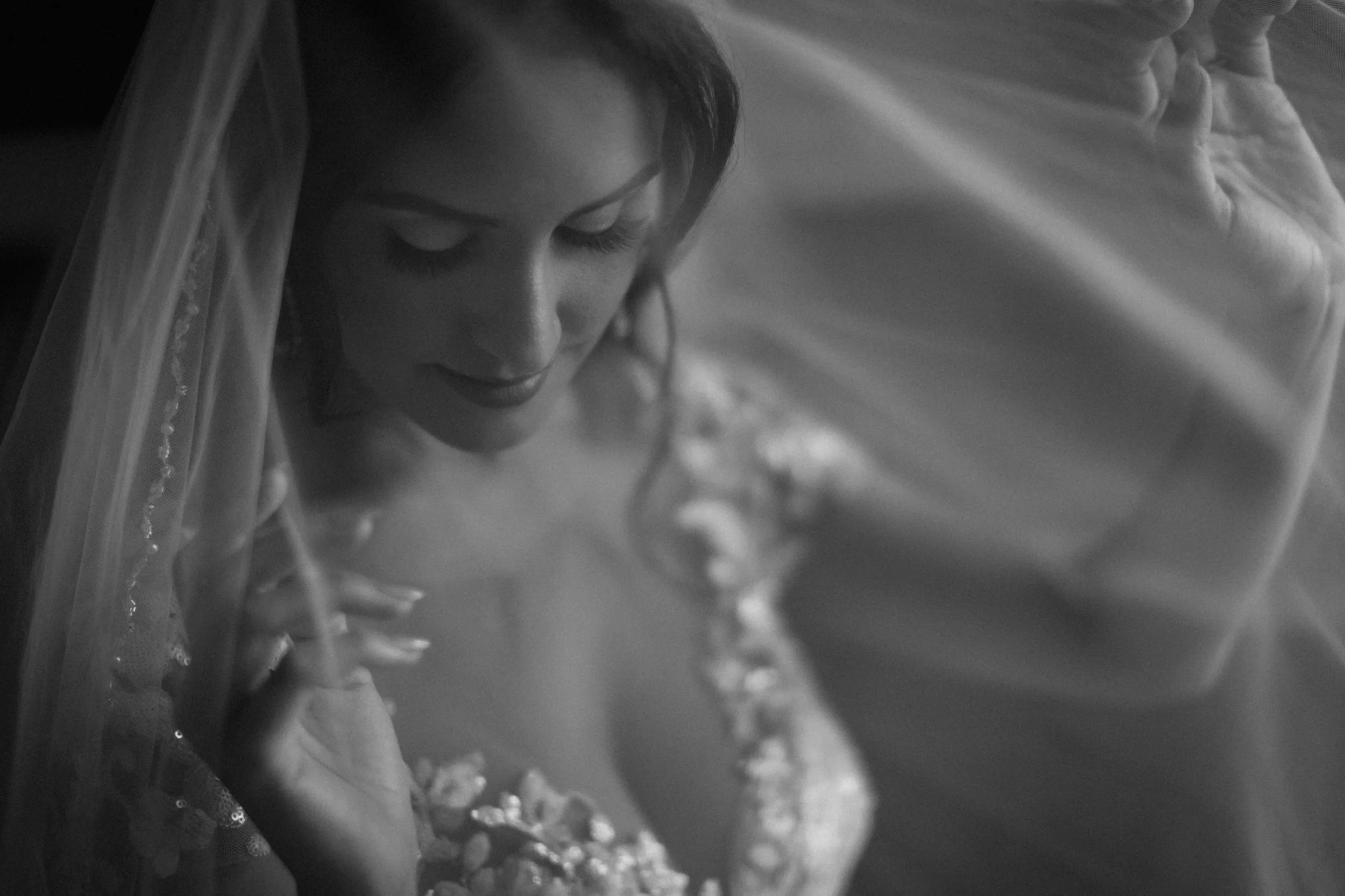A woman with dark hair and closed eyes, wearing a wedding dress and veil, is looking downward and touching her chest with one hand.
