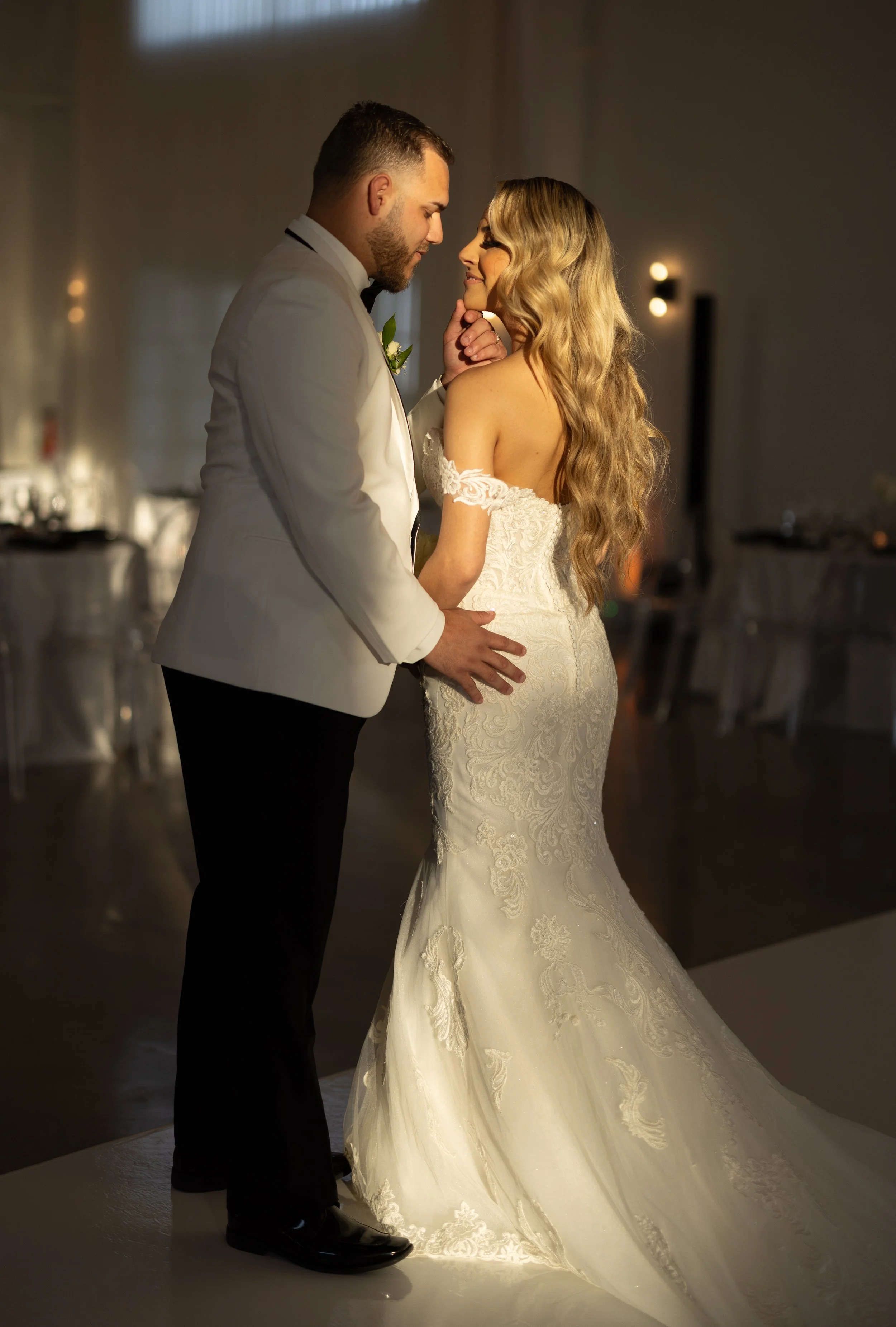 A bride and groom sharing a dance at their wedding reception. The groom wears a white tuxedo jacket and black pants, and the bride wears a white lace wedding gown with off-the-shoulder lace sleeves. They are gazing at each other tenderly in a dimly lit reception hall.