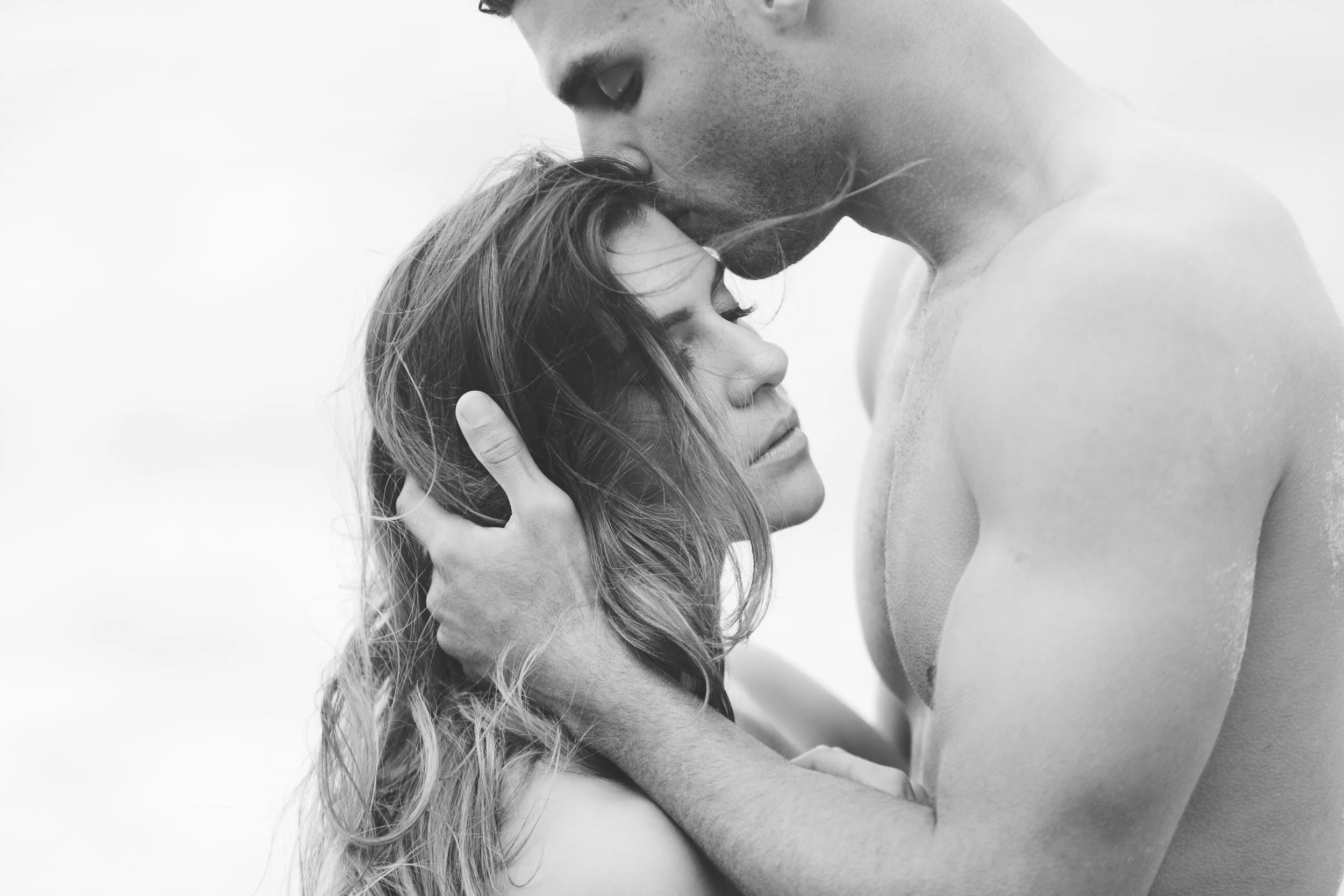 A black and white photograph of a man and woman in an intimate and emotional pose, with the man gently holding the woman's head and forearm, and their faces close together.