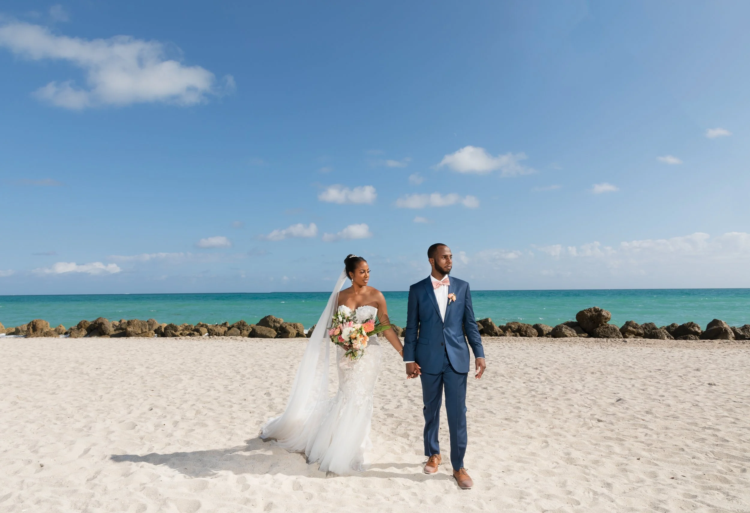A bride and groom holding hands walking on a sandy beach, with the ocean and blue sky with scattered clouds in the background.