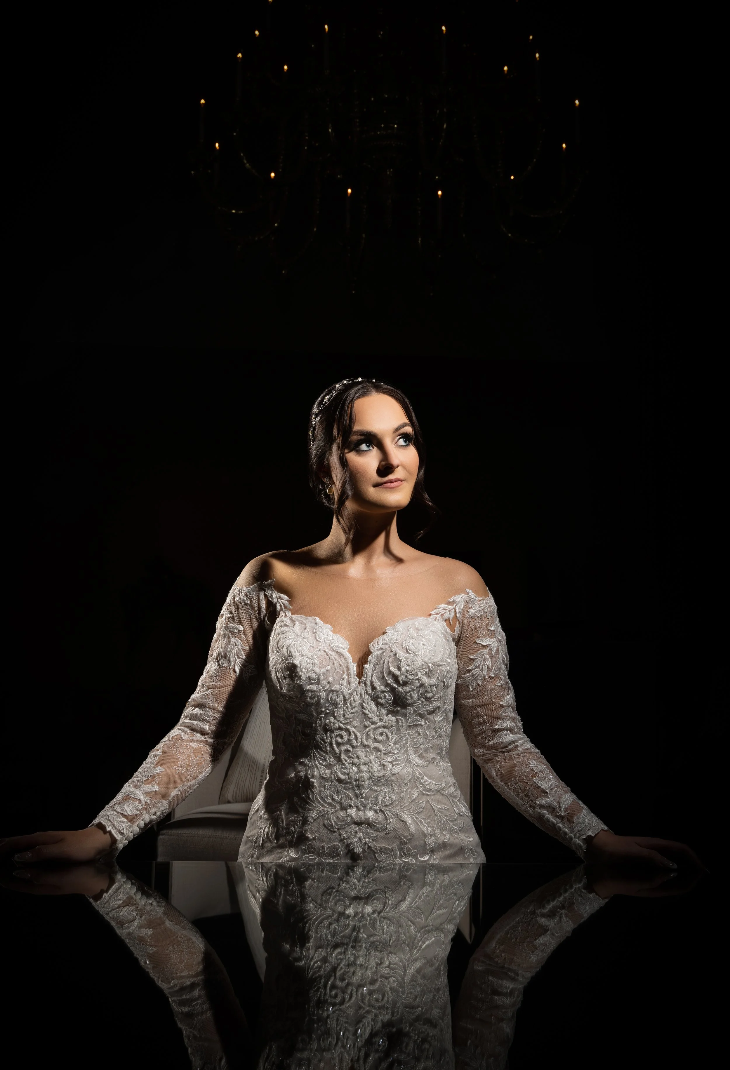 A bride in a white lace wedding dress with long sleeves, sitting at a reflective table with her hands resting on its surface. She has dark hair styled with a tiara and stands against a dark background with a chandelier overhead.