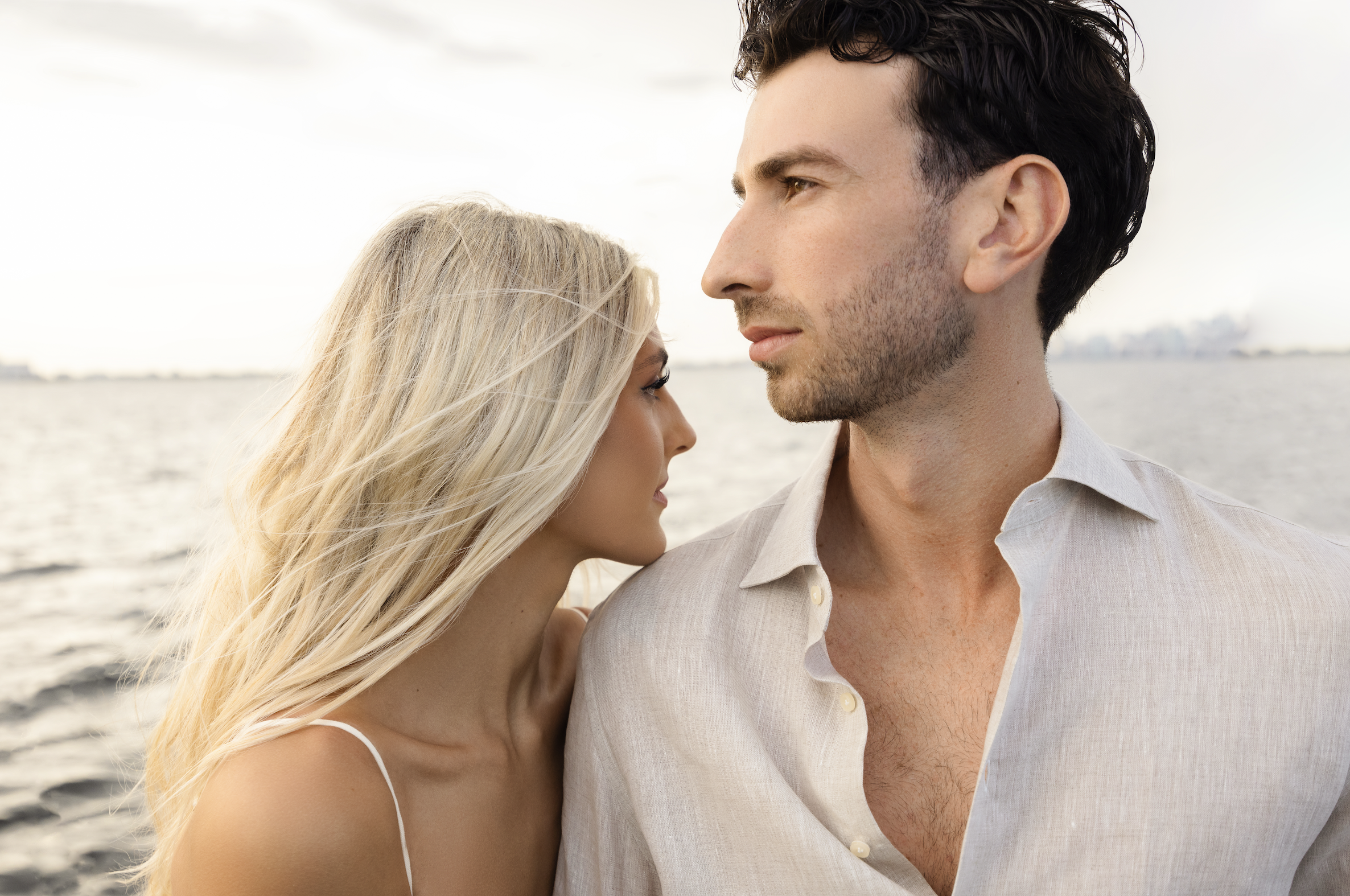 A man and woman near a body of water, facing each other closely, with the woman smiling and the man looking serious, during sunset.