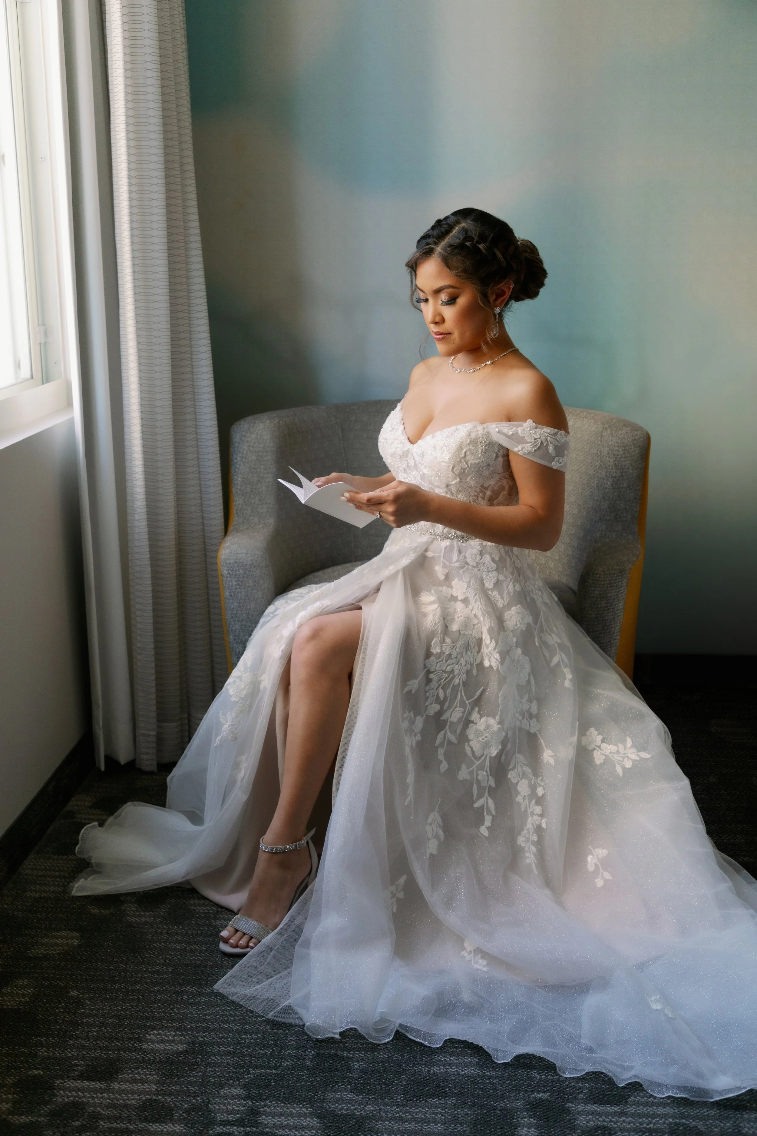 Bride sitting on a gray armchair by a window, reading a booklet, wearing a lace wedding gown with off-the-shoulder sleeves and a tulle skirt, silver high heels, and jewelry.
