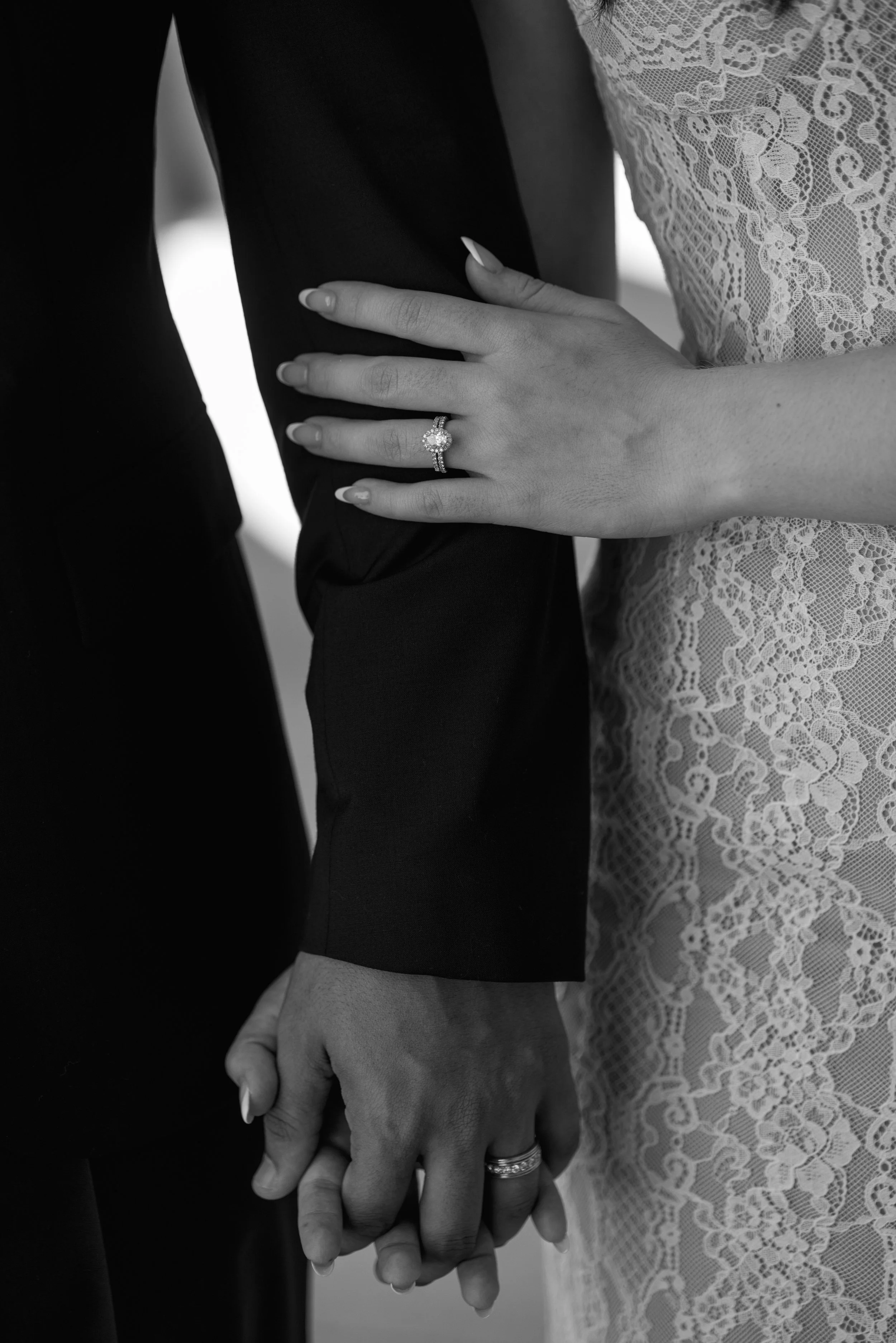 Close-up of a bride and groom holding hands, focusing on their wedding rings, with the bride's hand resting on the groom's arm. The bride is wearing a lace dress and an engagement ring, while the groom is in a black suit.