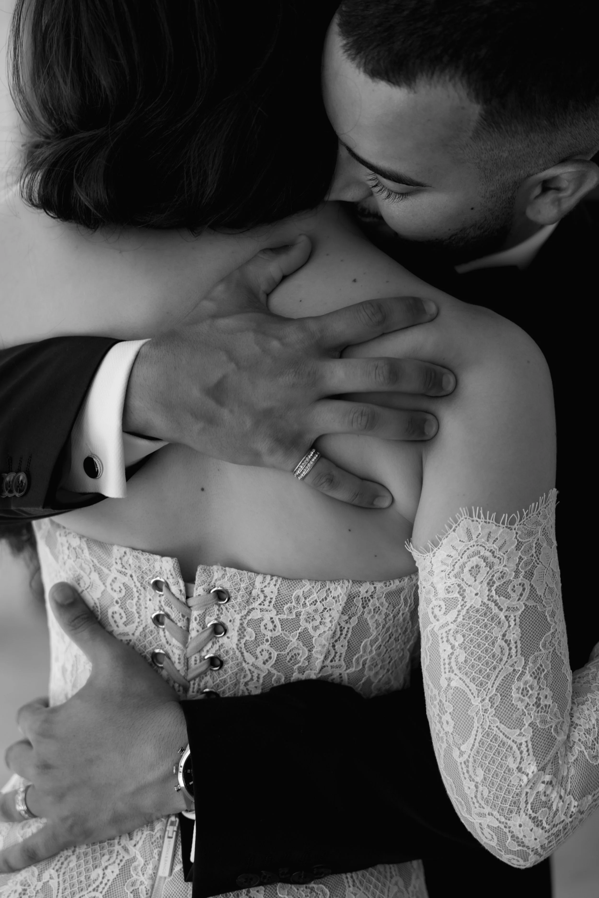 A close-up black and white photo of a couple embracing intensely, with the man kissing the woman’s shoulder. The woman is wearing a lace dress with cut-out details laced with a ribbon, and the man is dressed in a formal suit. The man’s hand is on her shoulder, showing a wedding ring, and her hand is on his arm, revealing a watch and another ring.