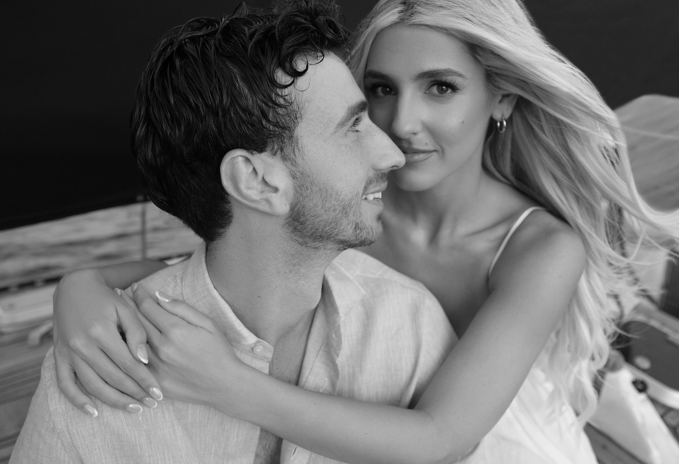 A black-and-white photograph of a smiling couple embracing, with the woman looking into the camera and the man looking at her, on a boat with water in the background.