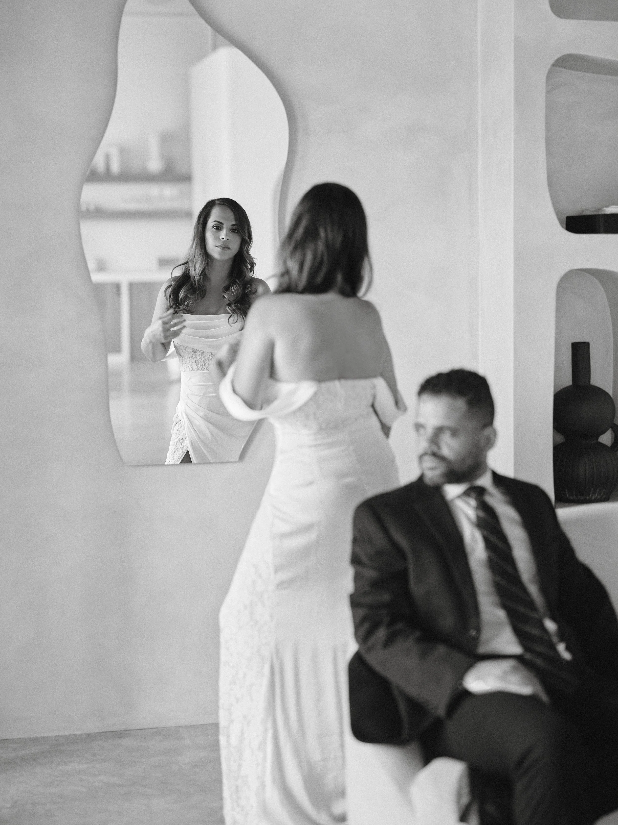 A woman looking into a mirror as she holds her hair, with a man sitting nearby in a modern room.