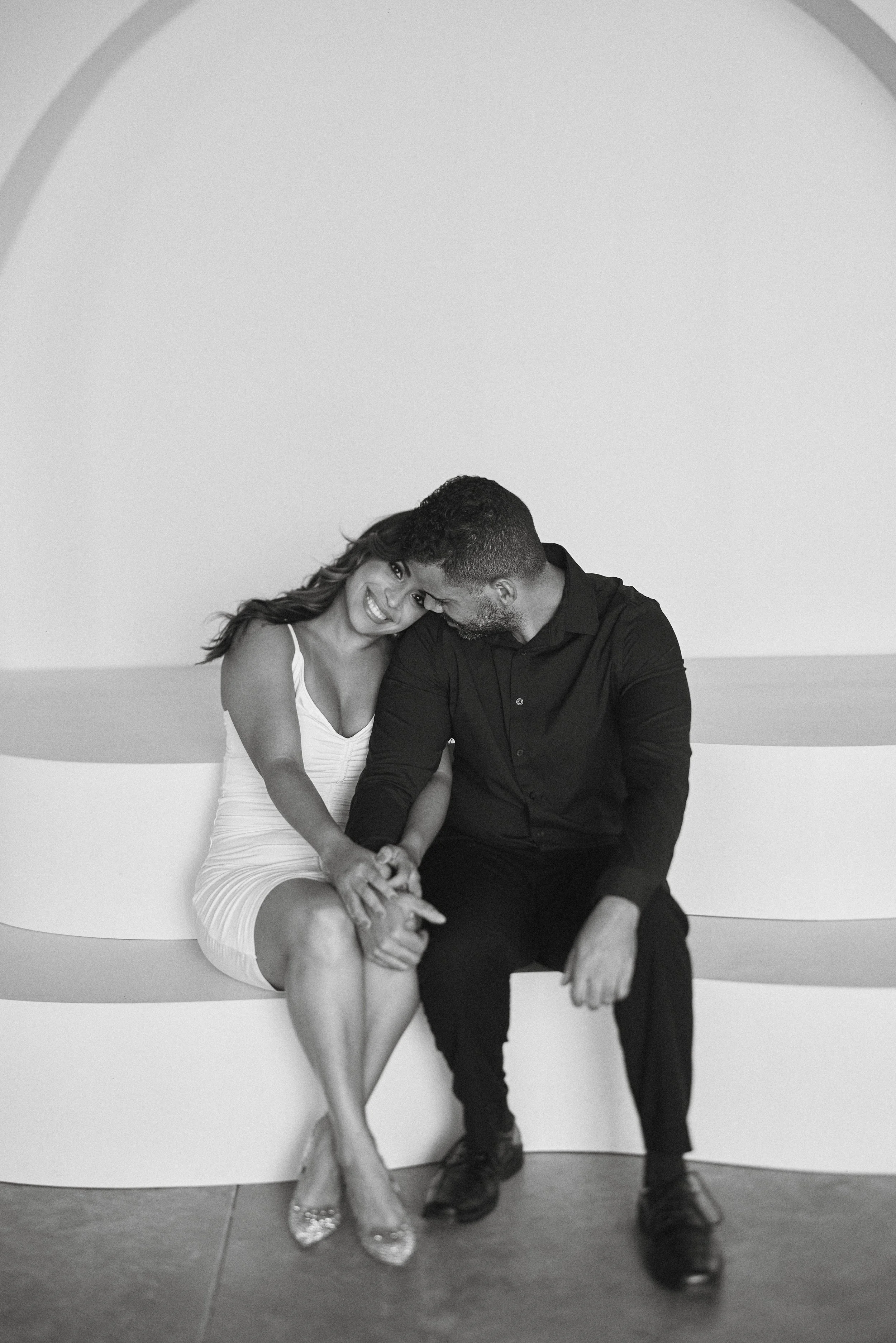 A black and white photo of a happy couple sitting close together, holding hands, smiling, and leaning their heads towards each other.