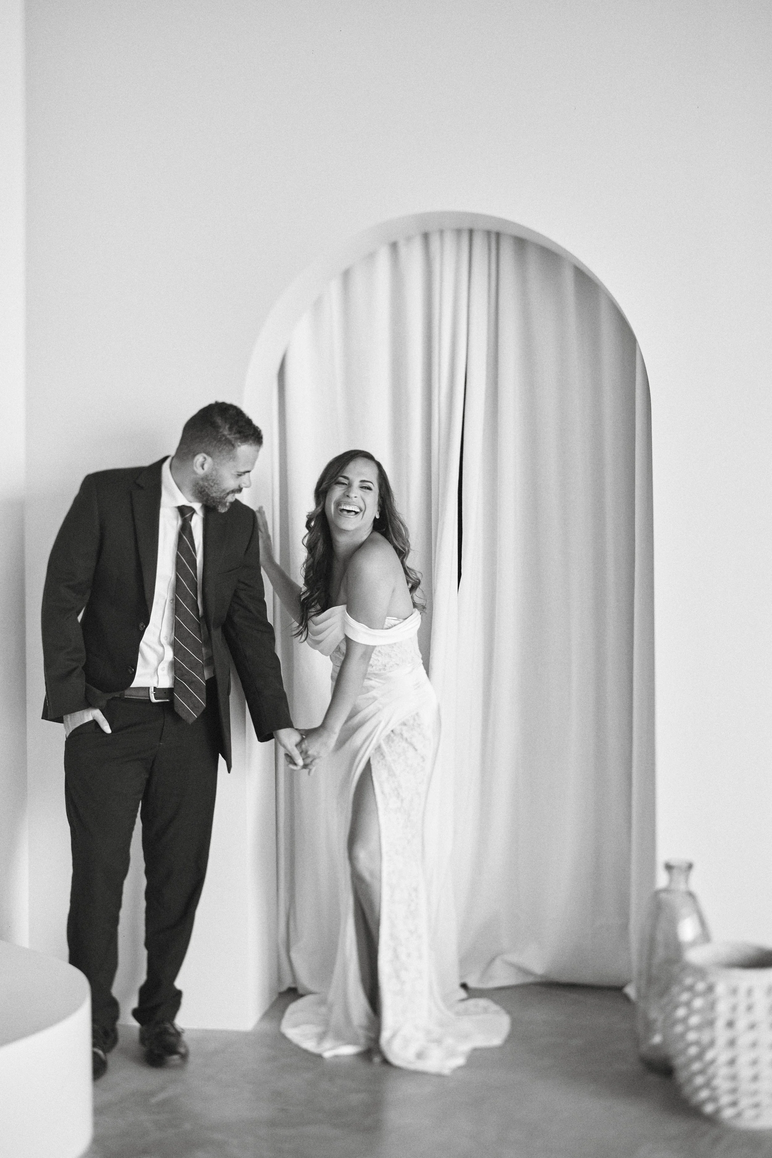 A joyful bride and groom sharing a moment in front of a curtain, holding hands and laughing.
