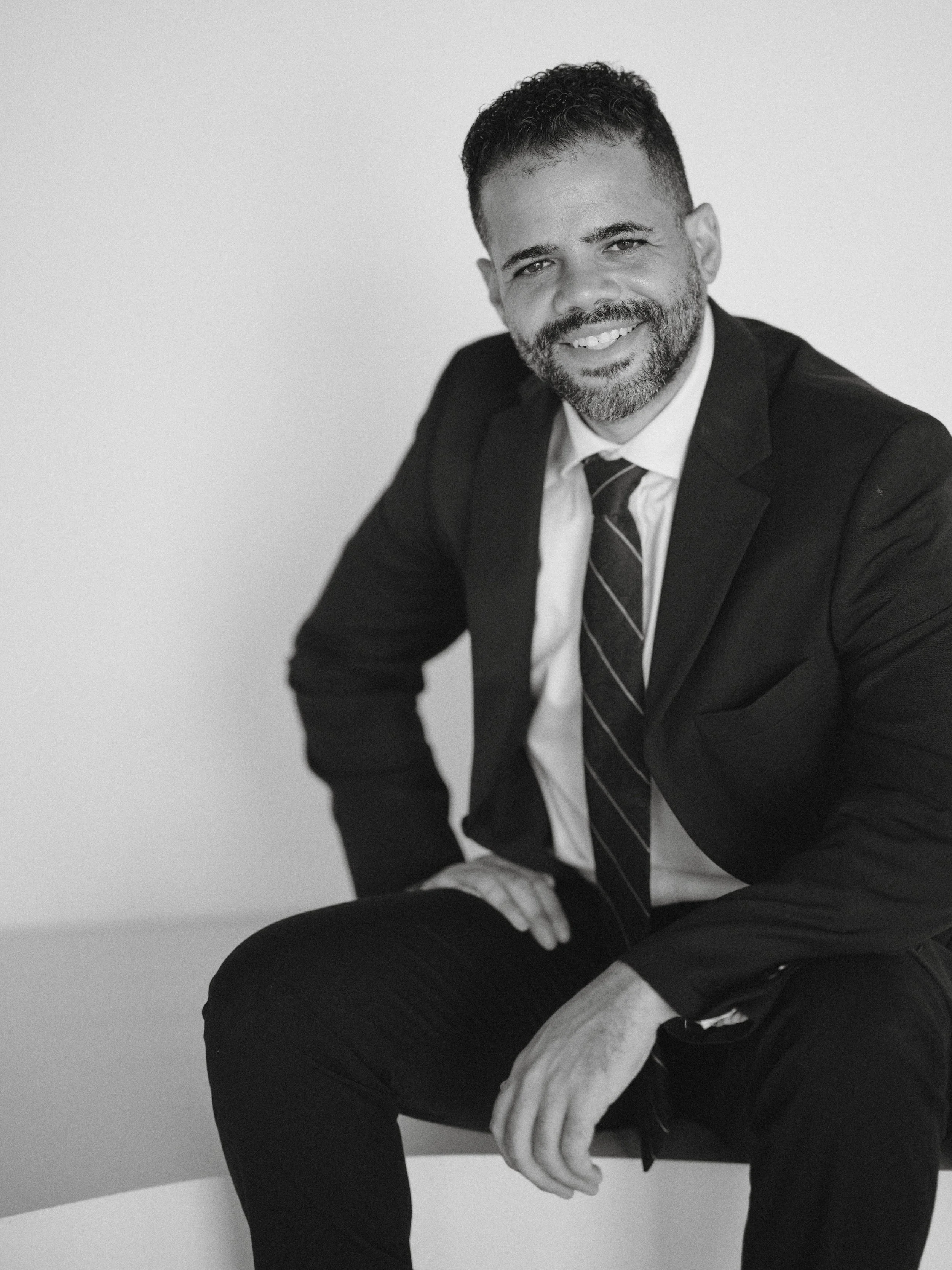 Black and white portrait of a man in a suit, smiling, sitting with one hand on his knee and the other on his thigh, against a plain background.