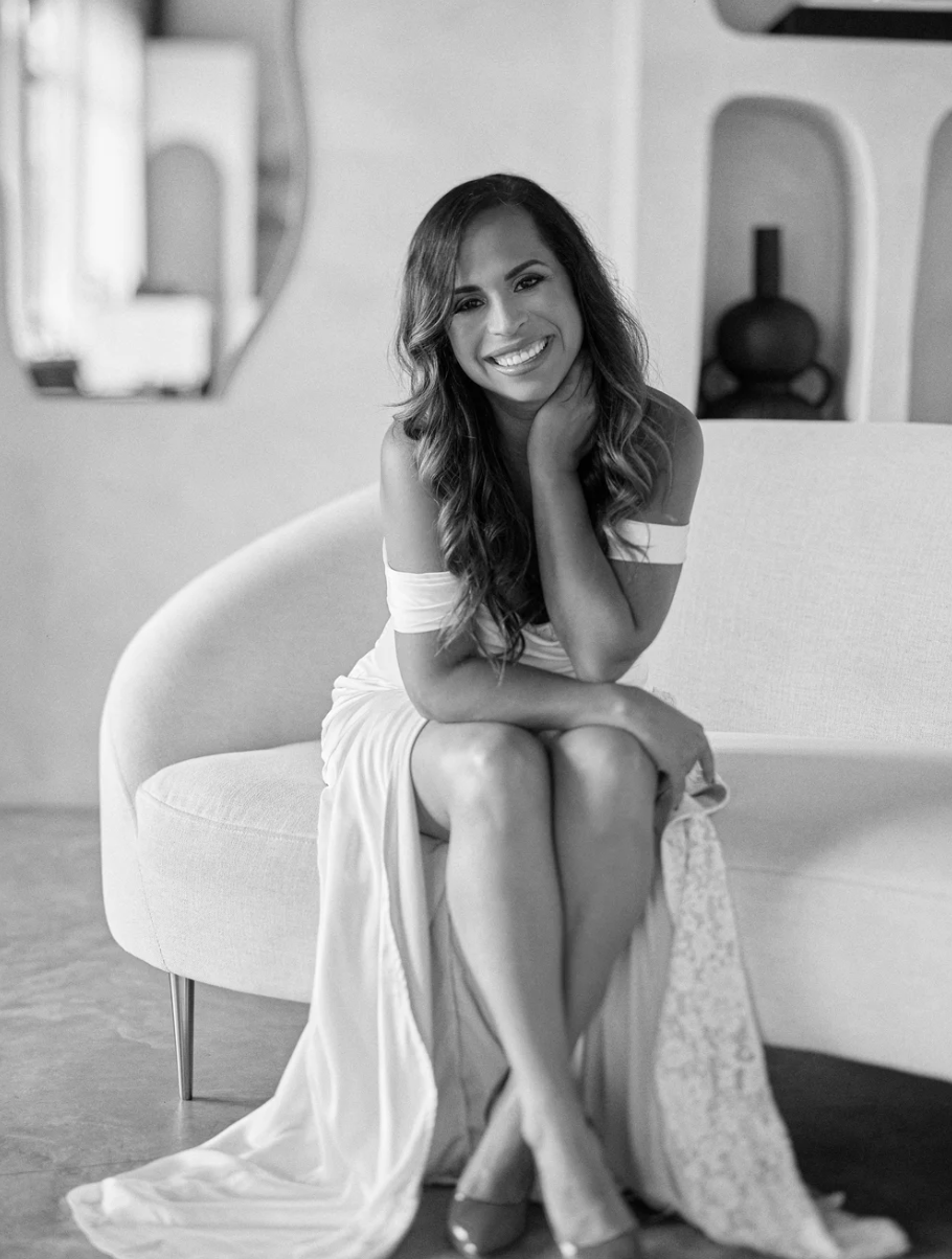 A smiling woman with long wavy hair, sitting on a modern sofa, in a bright living room, wearing an off-the-shoulder dress.