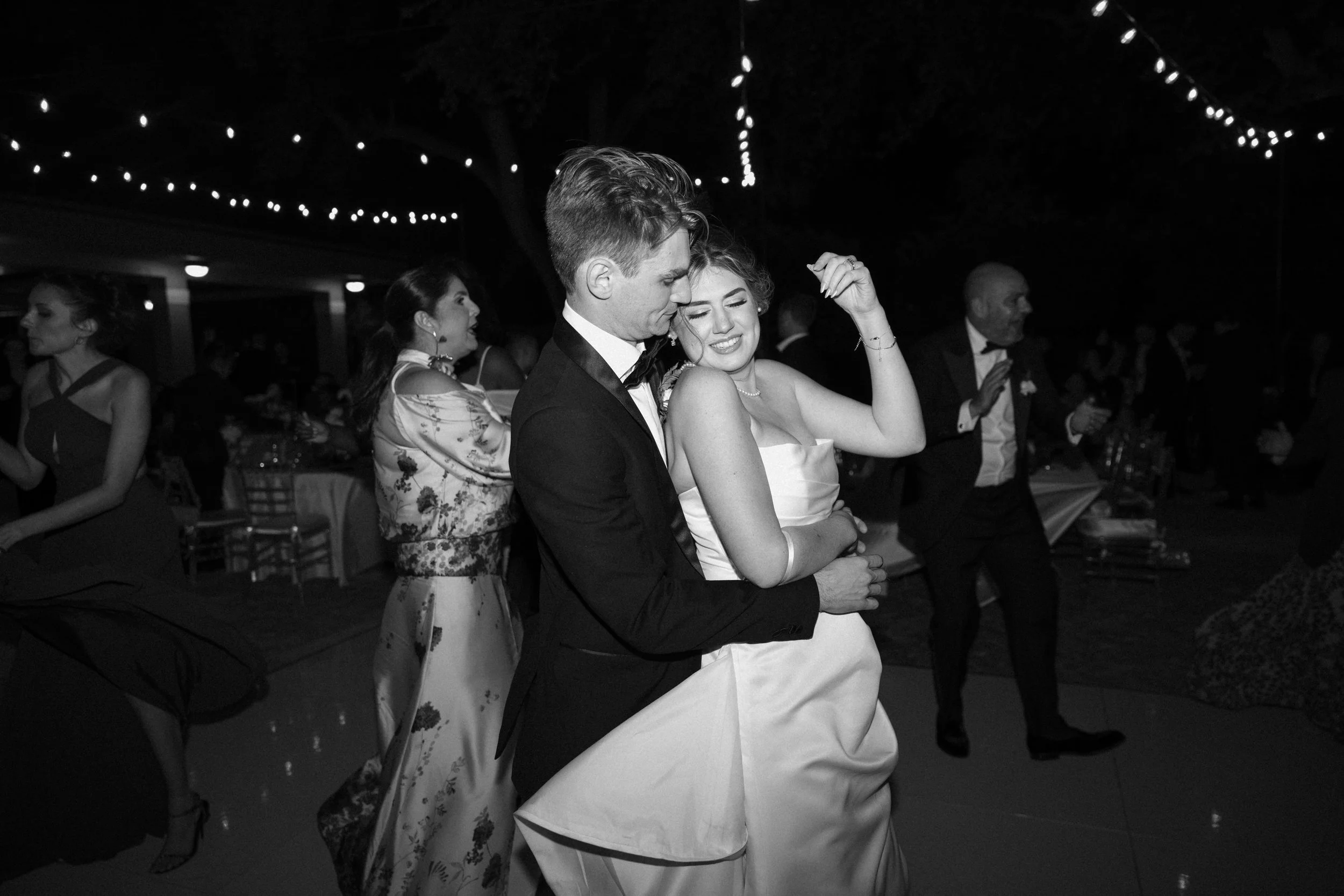 A black and white photo of a bride and groom dancing closely at their wedding reception, surrounded by guests under string lights.