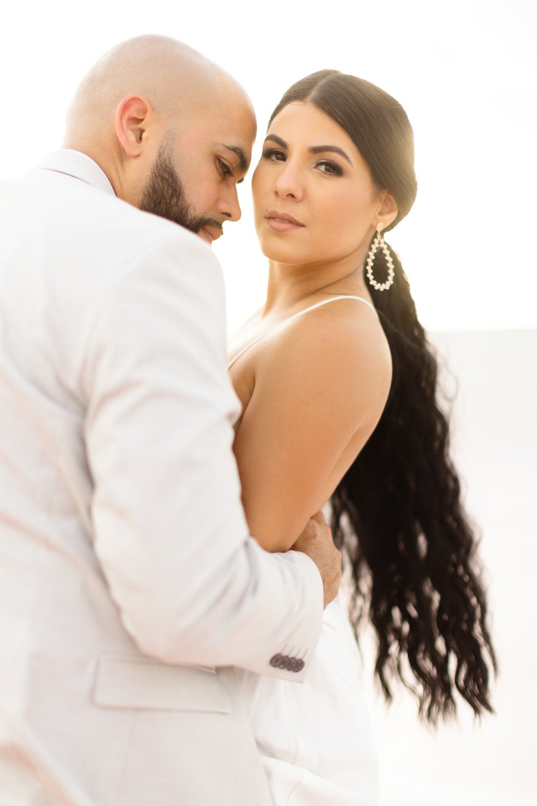 A couple dressed in white, with the woman having long dark hair and large earrings, and the man having a beard and a shaved head, standing close together in an intimate pose.