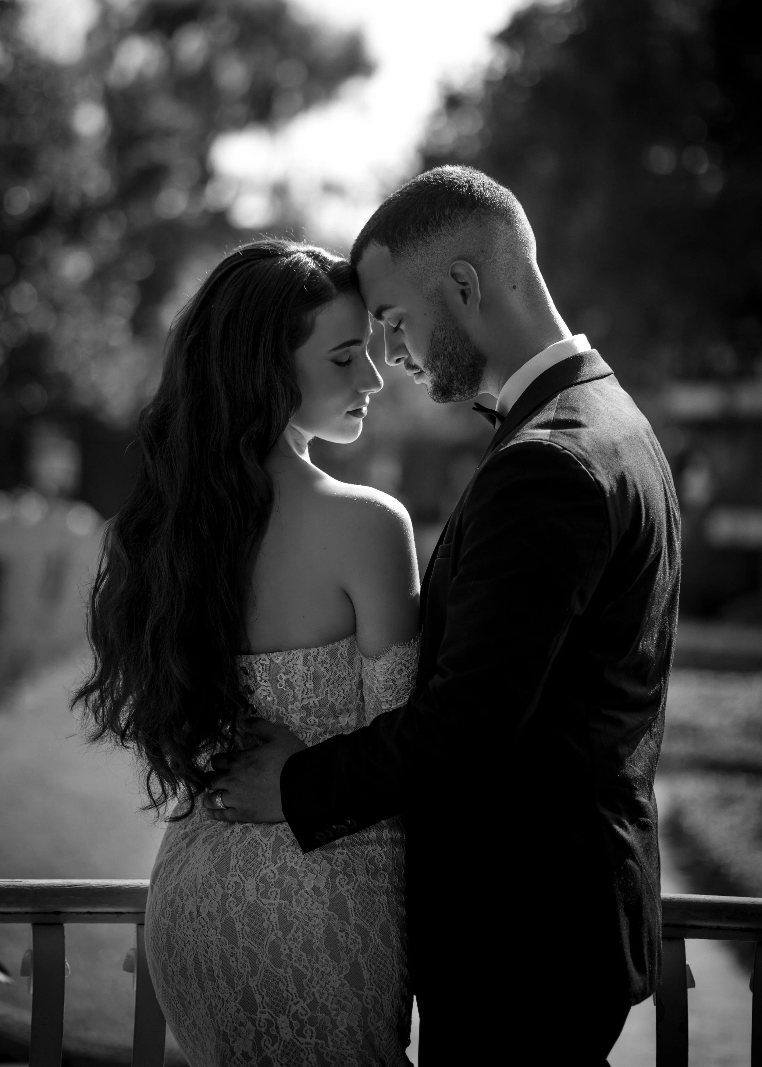 A black and white photo of a couple in wedding attire embracing, with foreheads touching, outdoors with blurred trees in the background.