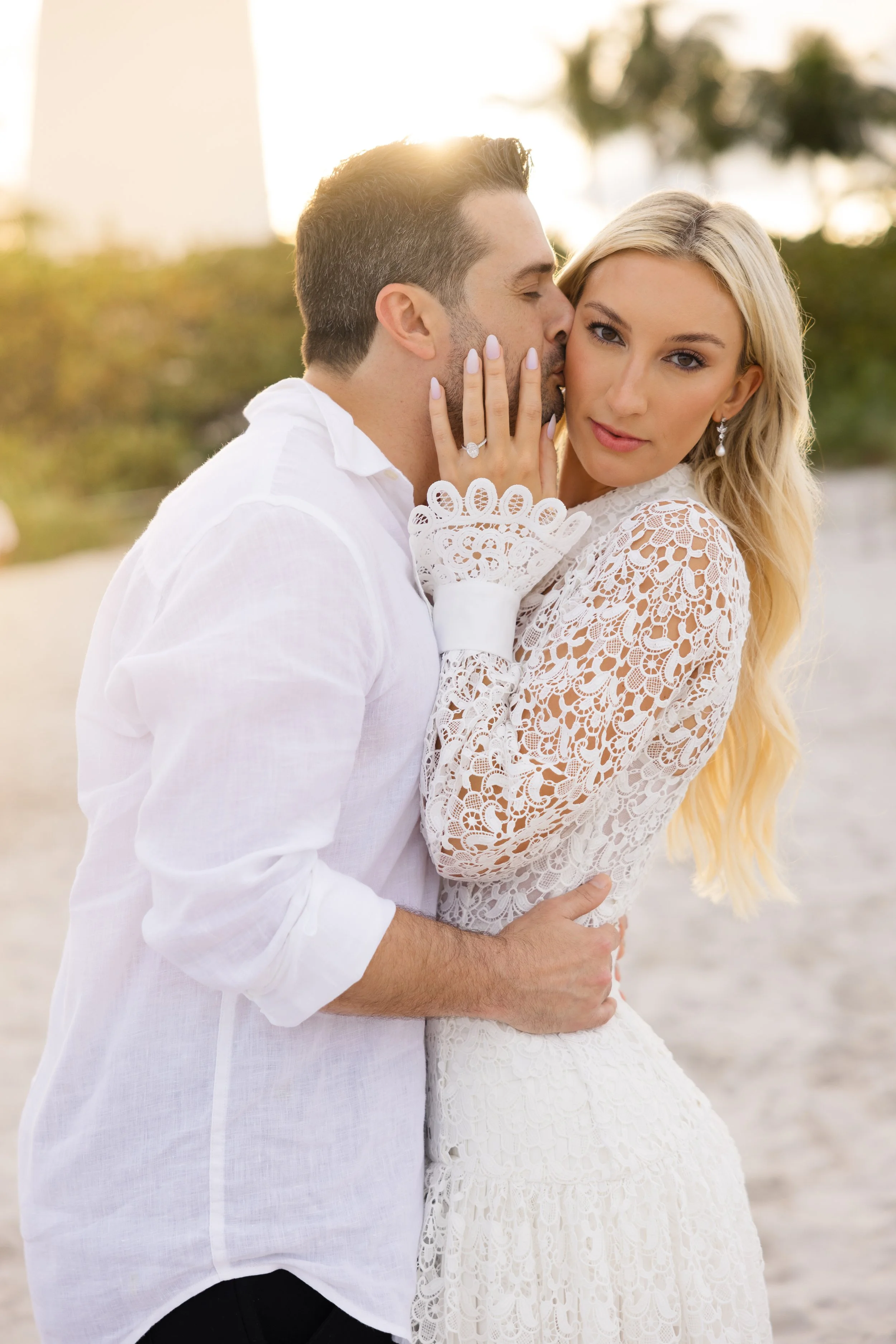 A couple dressed in white, with the man kissing the woman's cheek and the woman looking at the camera, on a beach at sunset.
