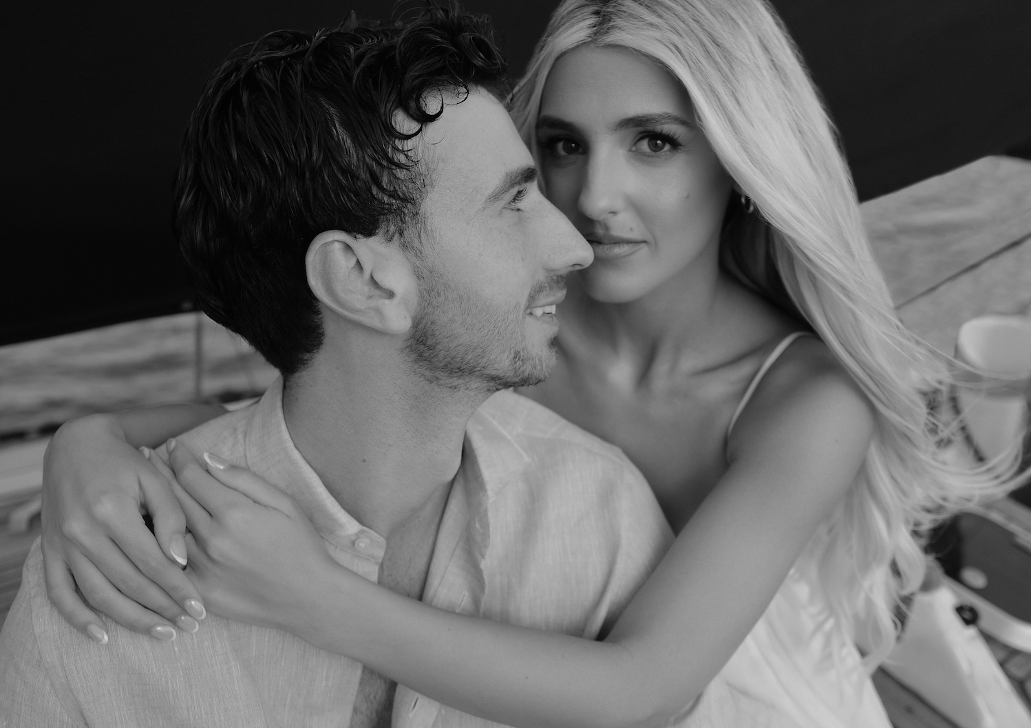 A black and white photo of a young couple, a man with dark wavy hair and a beard, and a woman with long, wavy blonde hair, embracing closely. The woman is looking directly at the camera while the man is focused on her, smiling softly. They are outdoors, sitting at a table with a beach and ocean in the background.