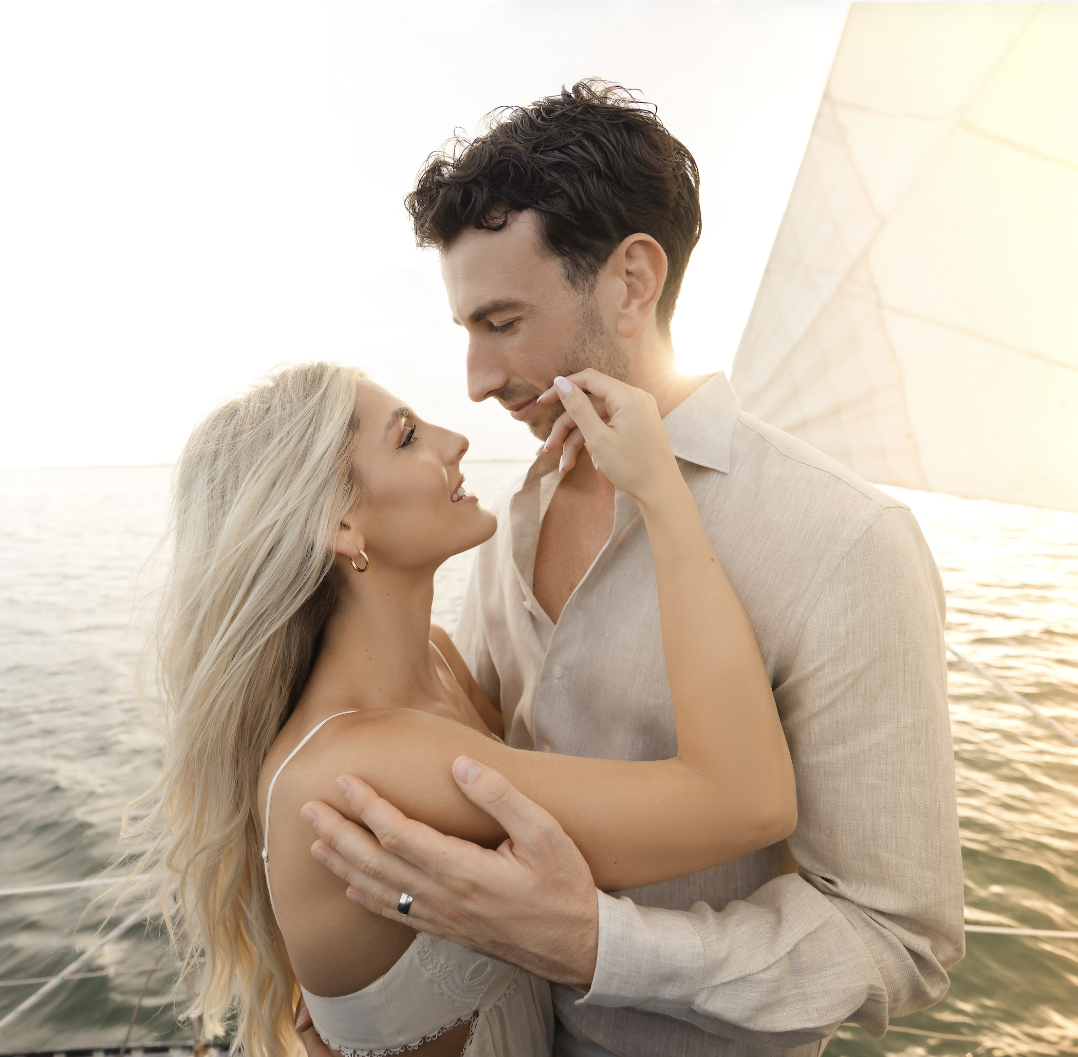 A couple with light skin and blonde hair embraces on a boat or near water at sunset, gazing into each other's eyes.