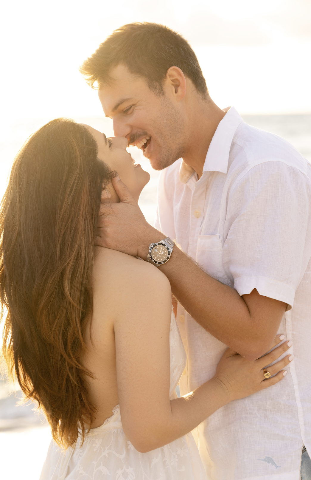A couple embracing on the beach during sunset, with the man gently holding the woman's face and both smiling intimately.