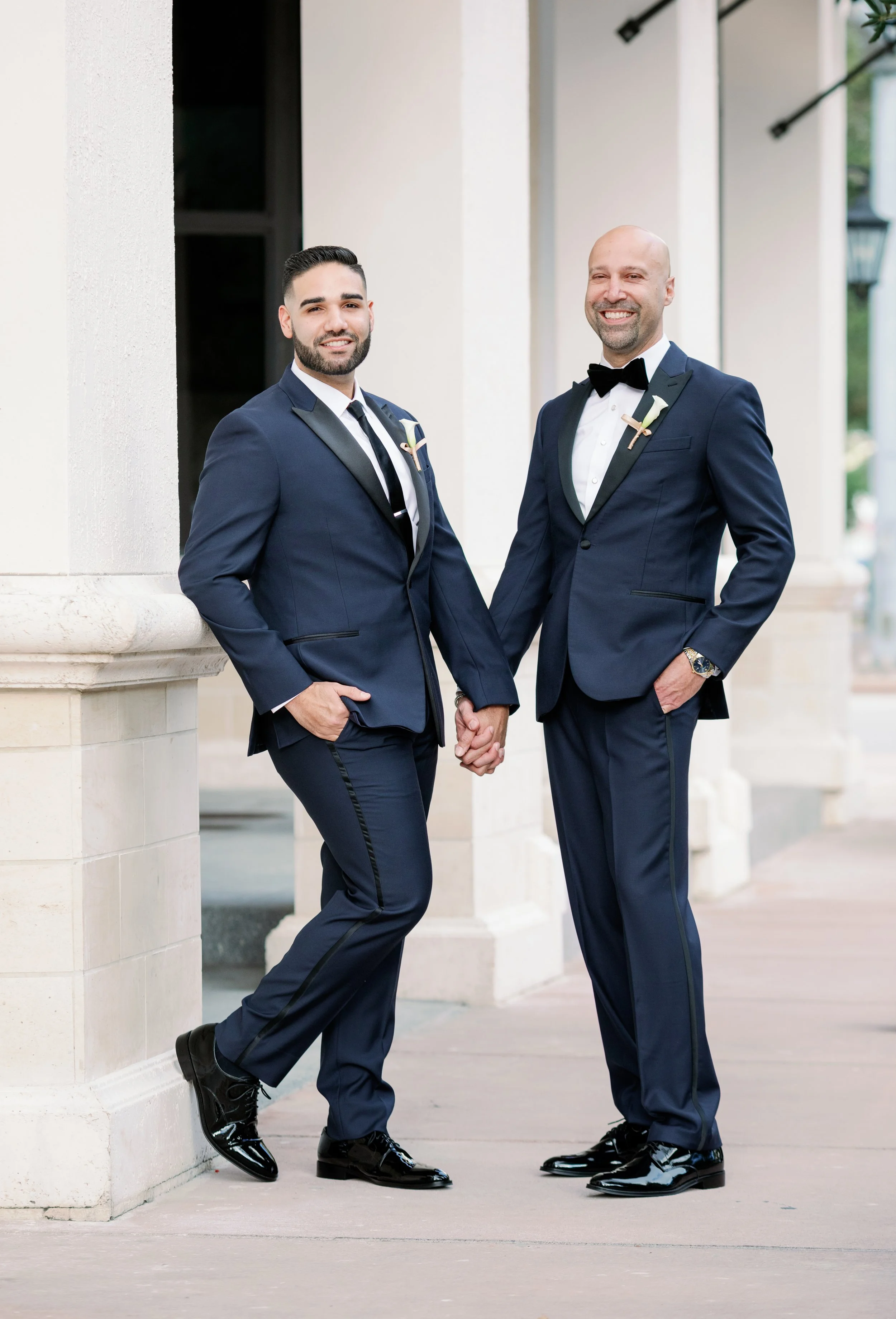 Two men in matching dark blue tuxedos holding hands and smiling outdoors near a building with white columns.