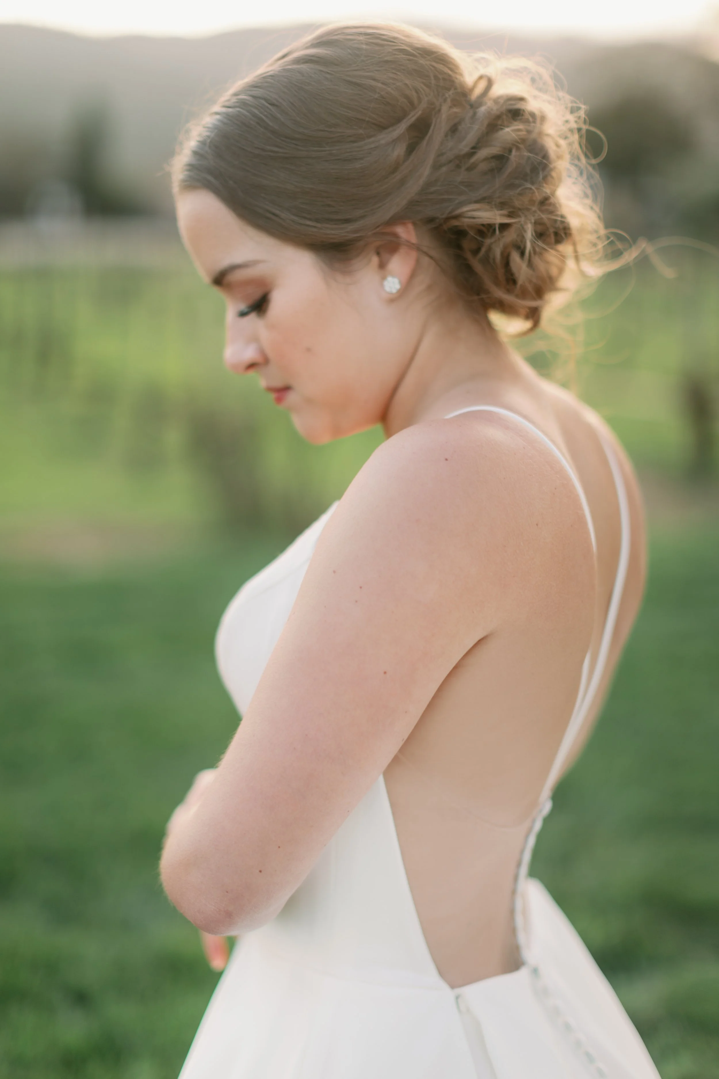 Close-up side view of a bride in a white wedding dress outdoors, with her head bowed and eyes closed, against a blurred green landscape background.