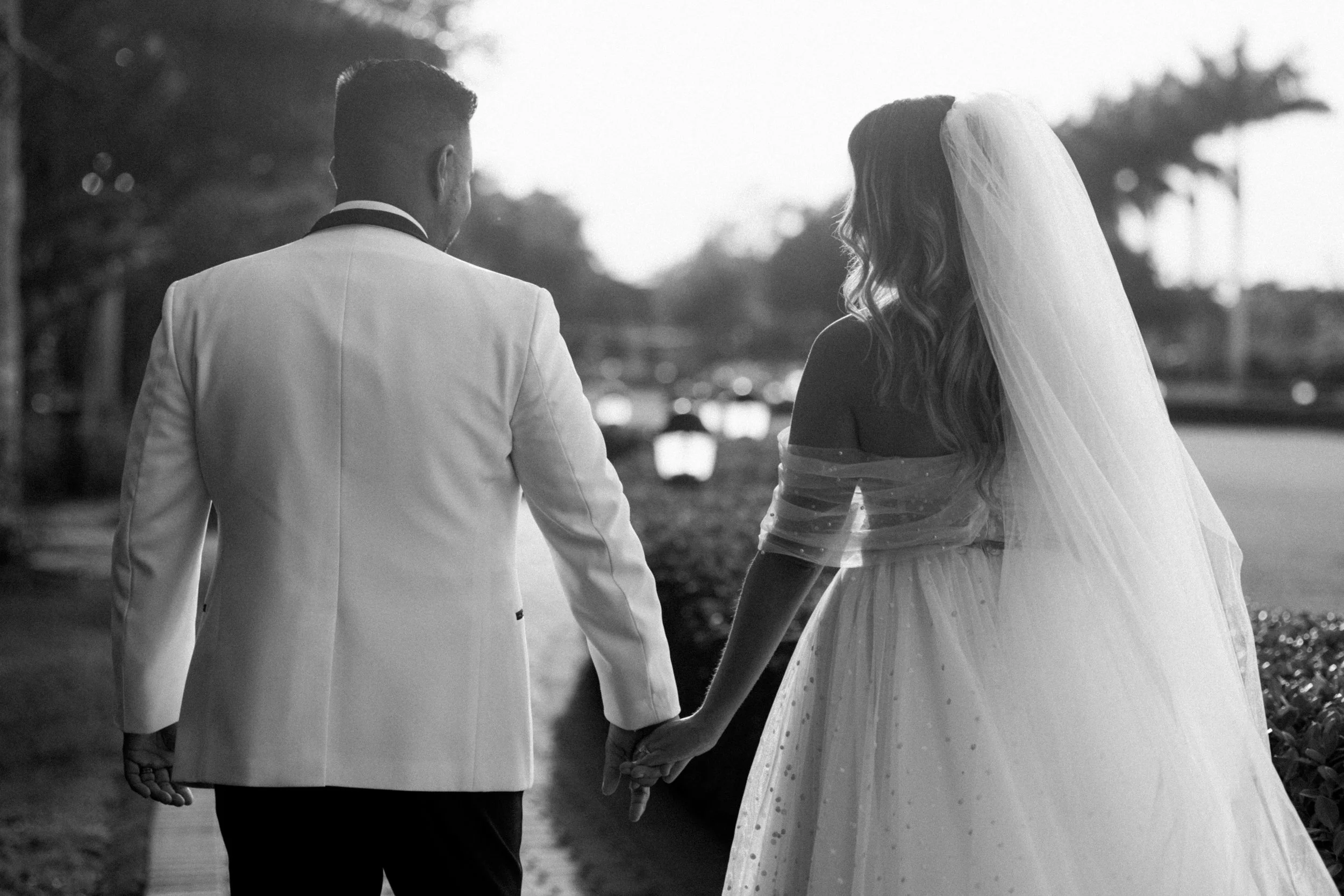 A black and white photo of a bride and groom holding hands and walking outdoors, viewed from behind.