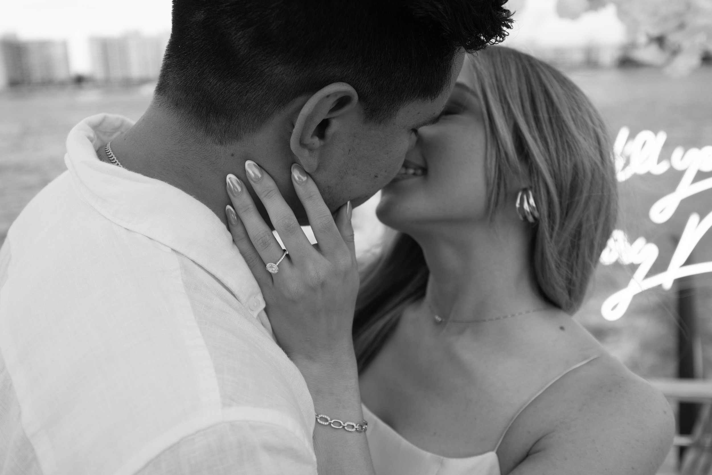 A black-and-white photo of a couple sharing a kiss. The woman has long hair and is wearing a ring, bracelet, earrings, and a sleeveless top. The man has short hair and is wearing a light-colored shirt, with his hand gently touching her face.