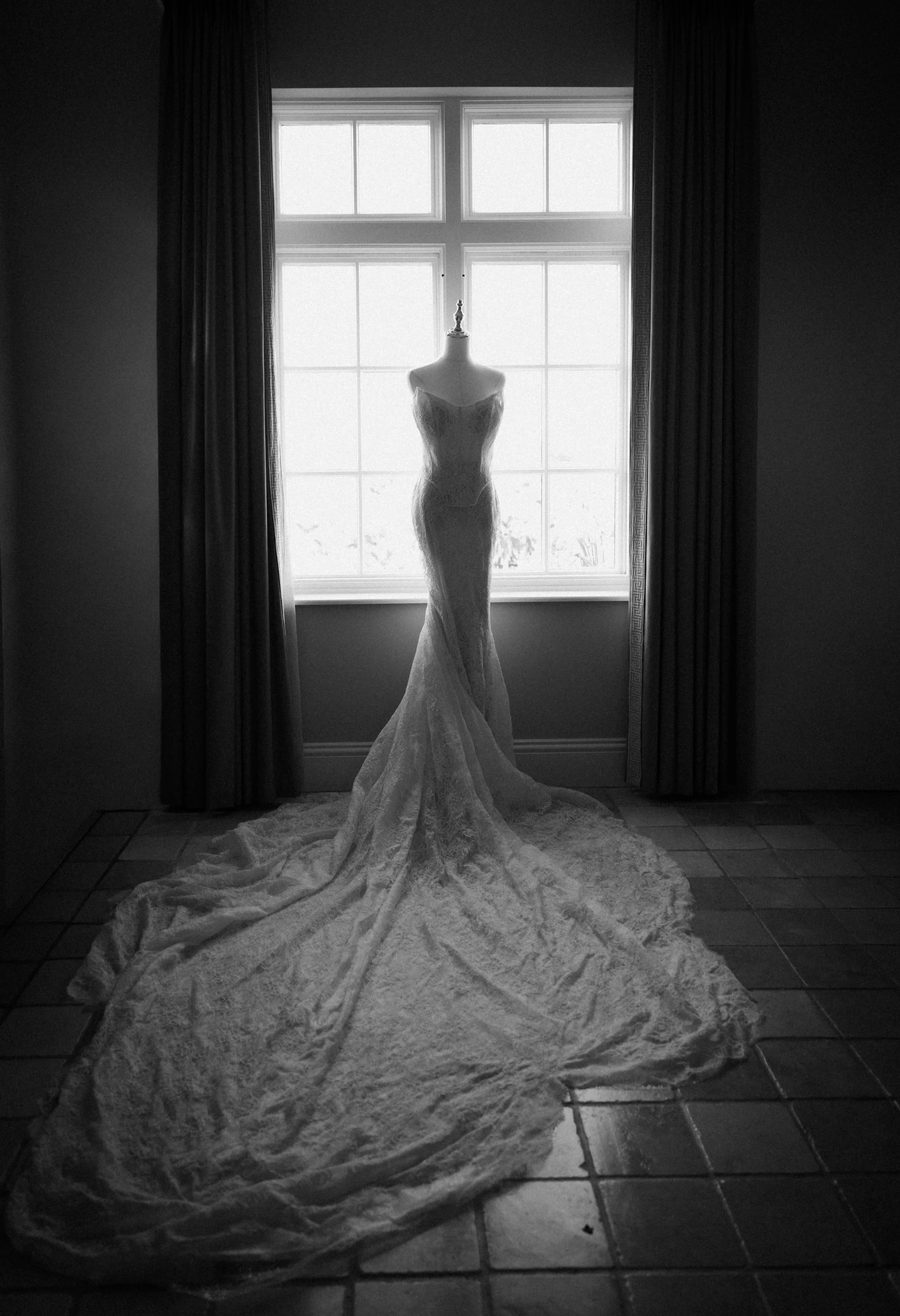 A wedding dress on a mannequin placed in front of a large window with curtains, in a room with tiled floor, illuminated from behind through the window.