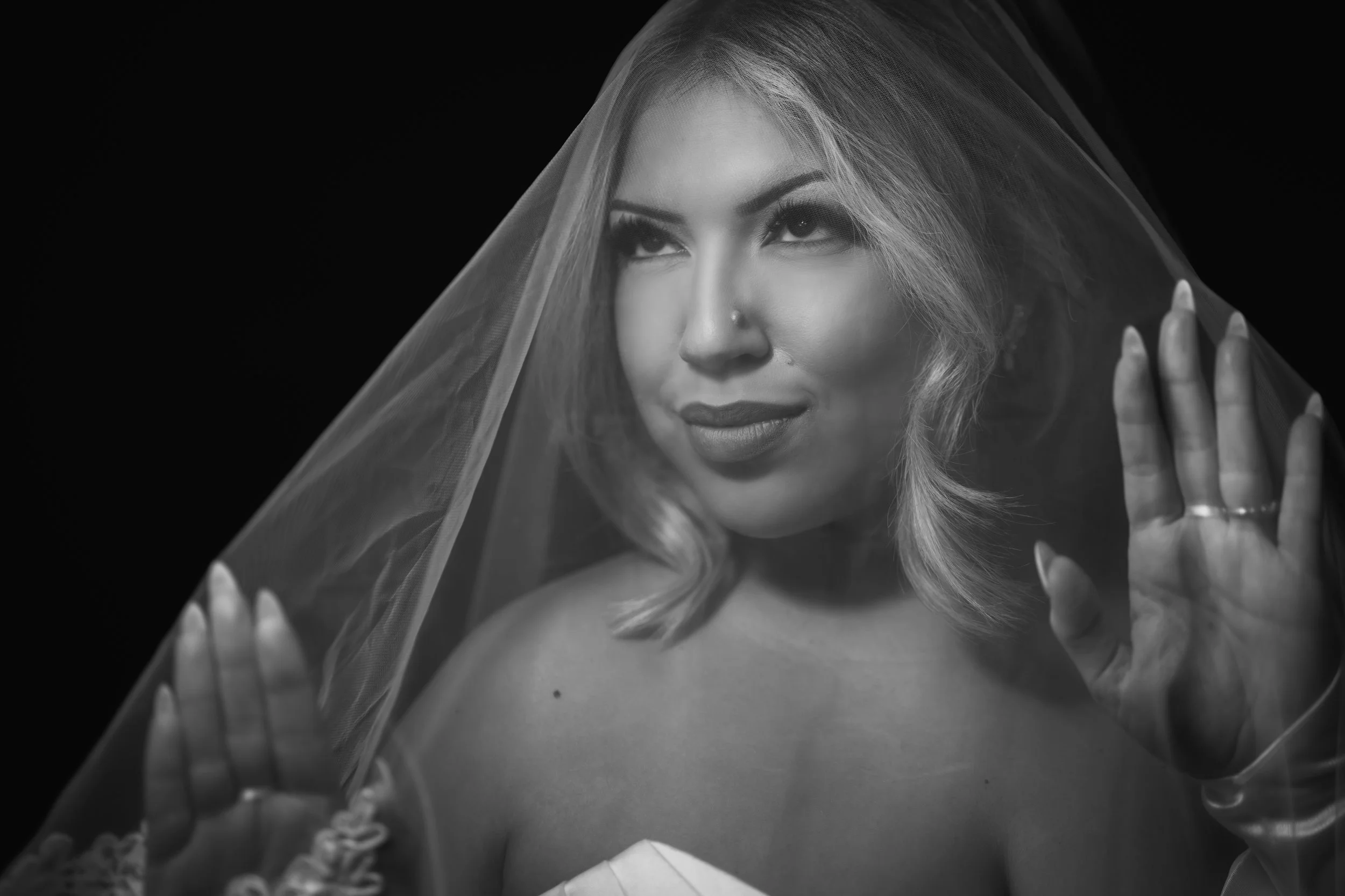 Black and white portrait of a woman with shoulder-length wavy hair, wearing a veil and minimal makeup, looking upward with a slight smile, holding the veil with both hands.