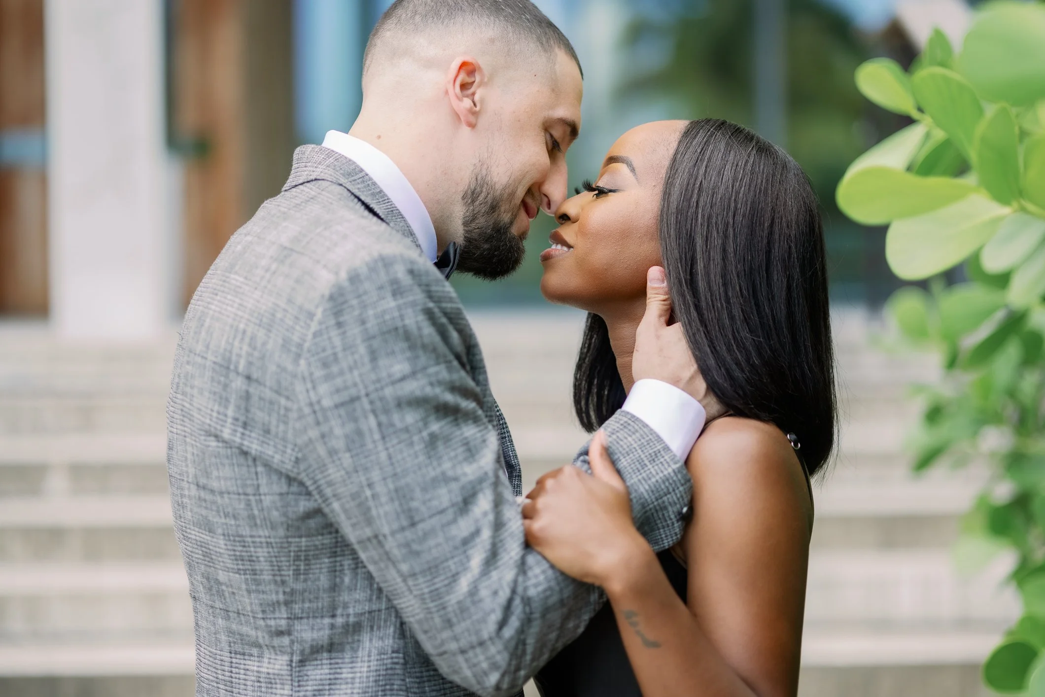 A couple is about to kiss, with their faces close and eyes closed. The man has a beard and is wearing a gray checked suit with a white shirt and bow tie. The woman has straight black hair, makeup, and is wearing a black sleeveless dress, holding the 