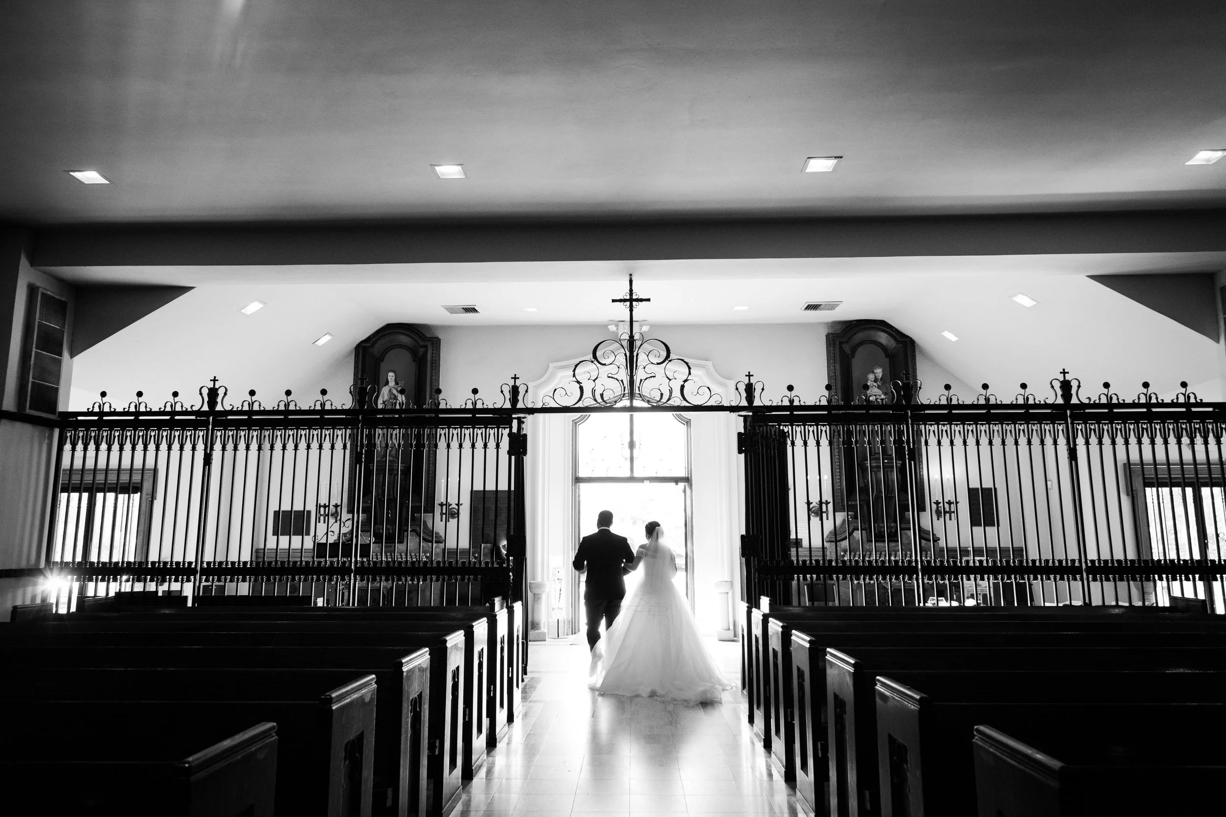 A bride and groom walking arm in arm out of a church, silhouetted against the bright outdoor light, with pews and religious statues visible inside.