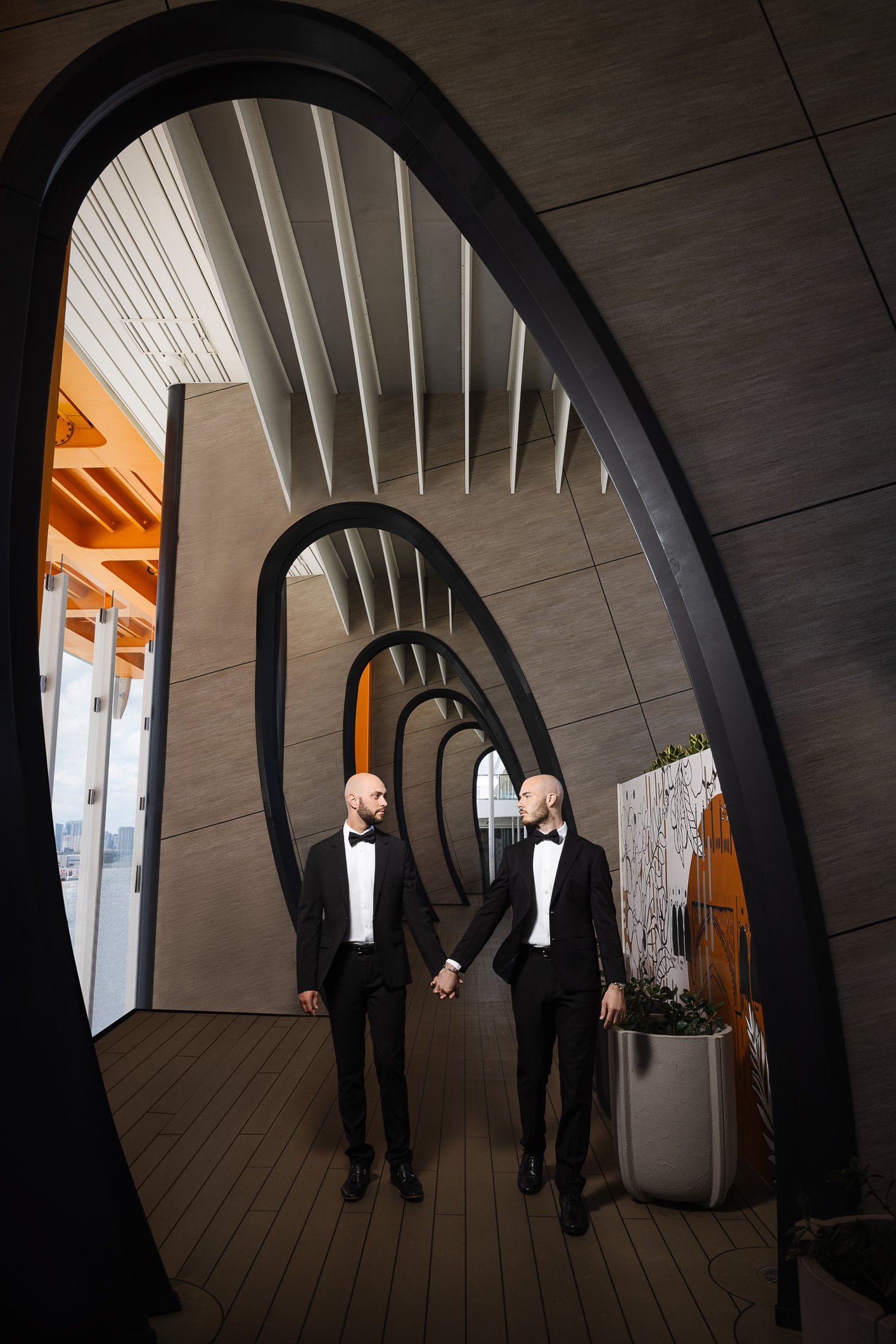 Two men in black tuxedos holding hands and walking through a modern hallway with a spiral mirror reflection.