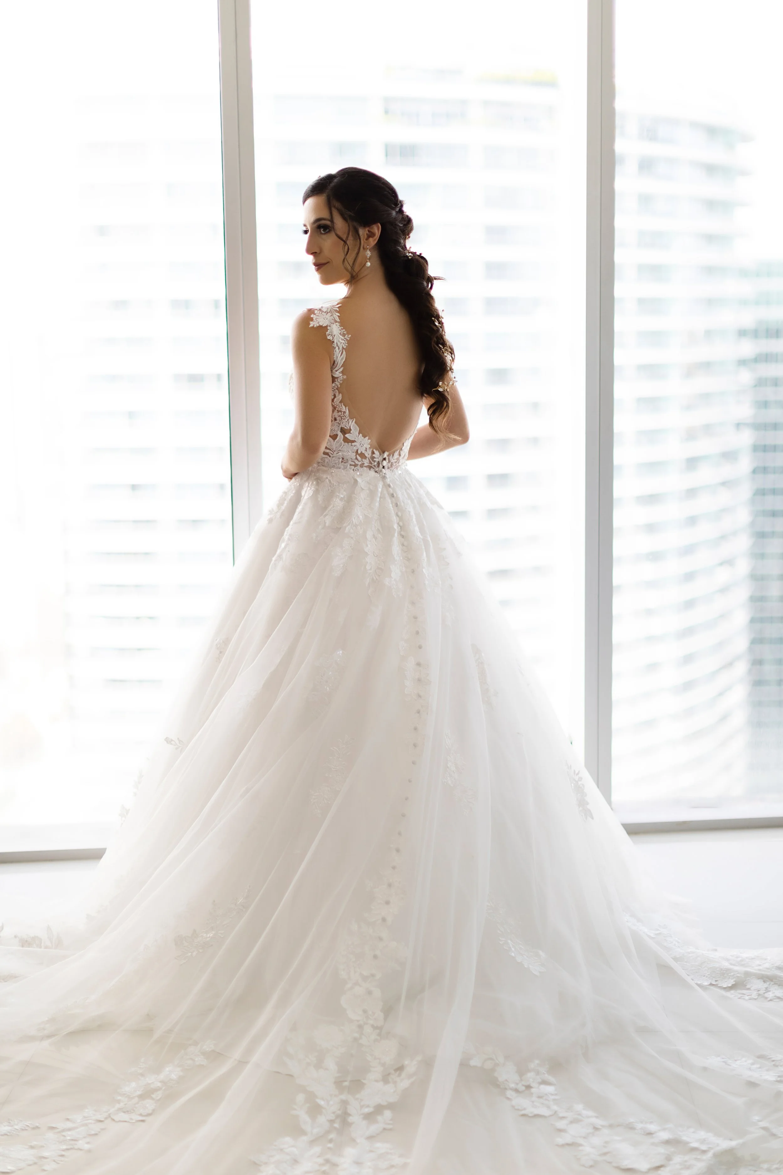 A bride in an elegant wedding gown with a low back, lace details, and a long trailing train, standing by a large window with city buildings outside.