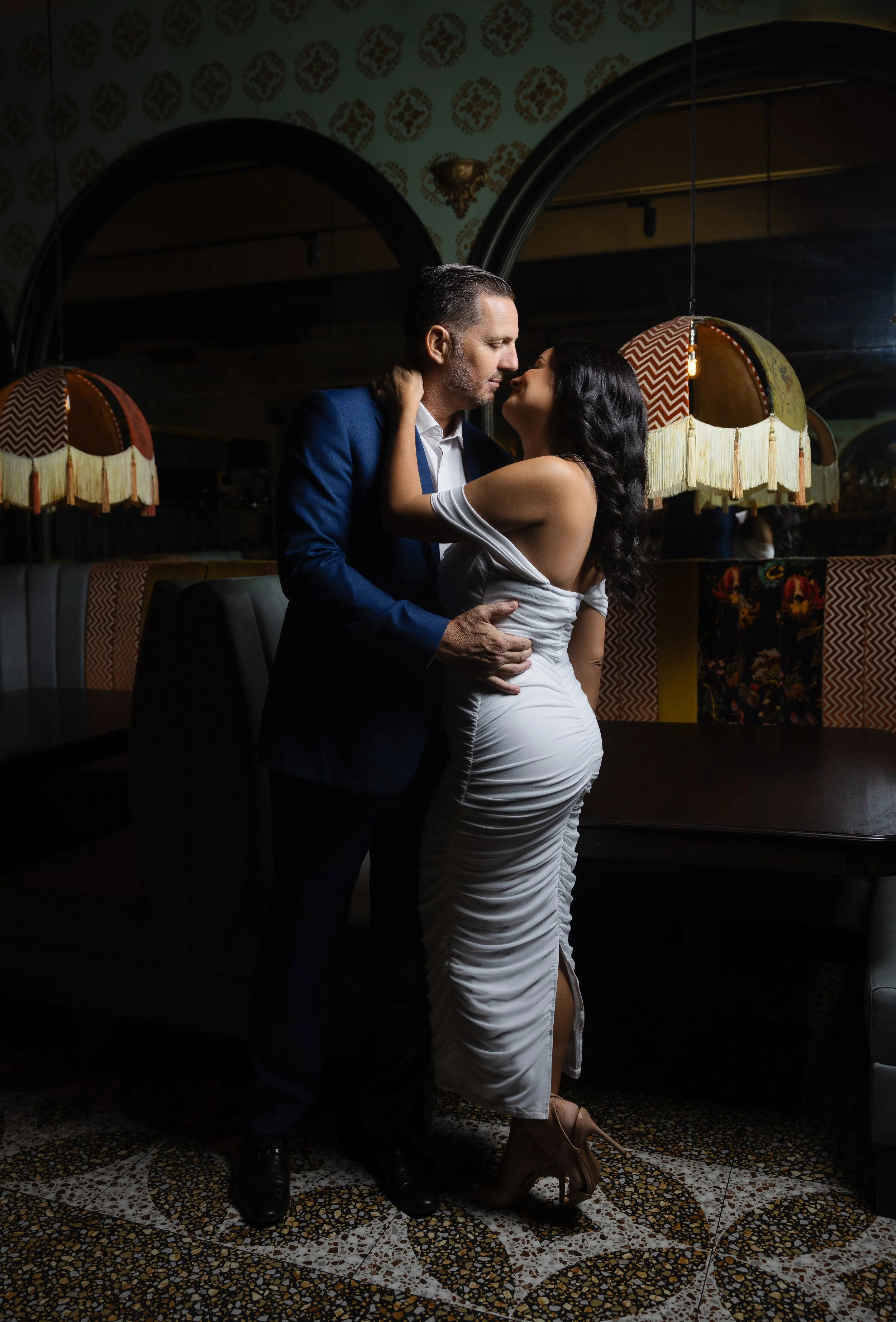 A romantic couple in an intimate embrace at a dimly lit restaurant or bar, with decorative lamps, patterned walls, and a dark wooden table in the background.