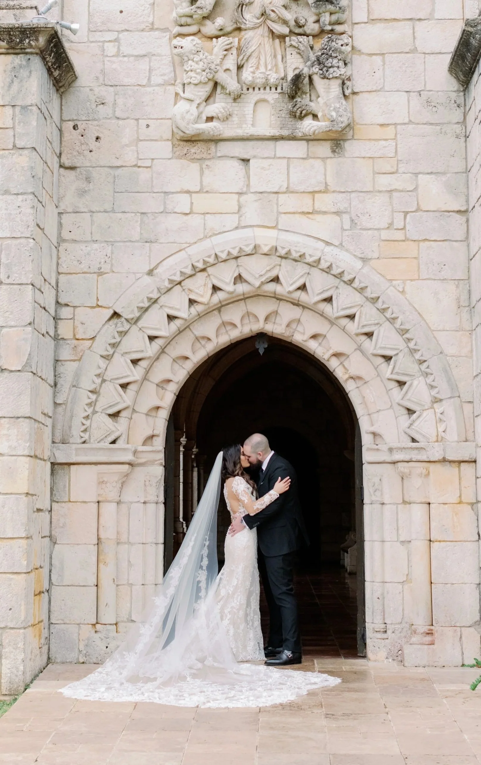 A bride and groom kissing in front of a historic stone church archway, with a decorative stone shield above them.