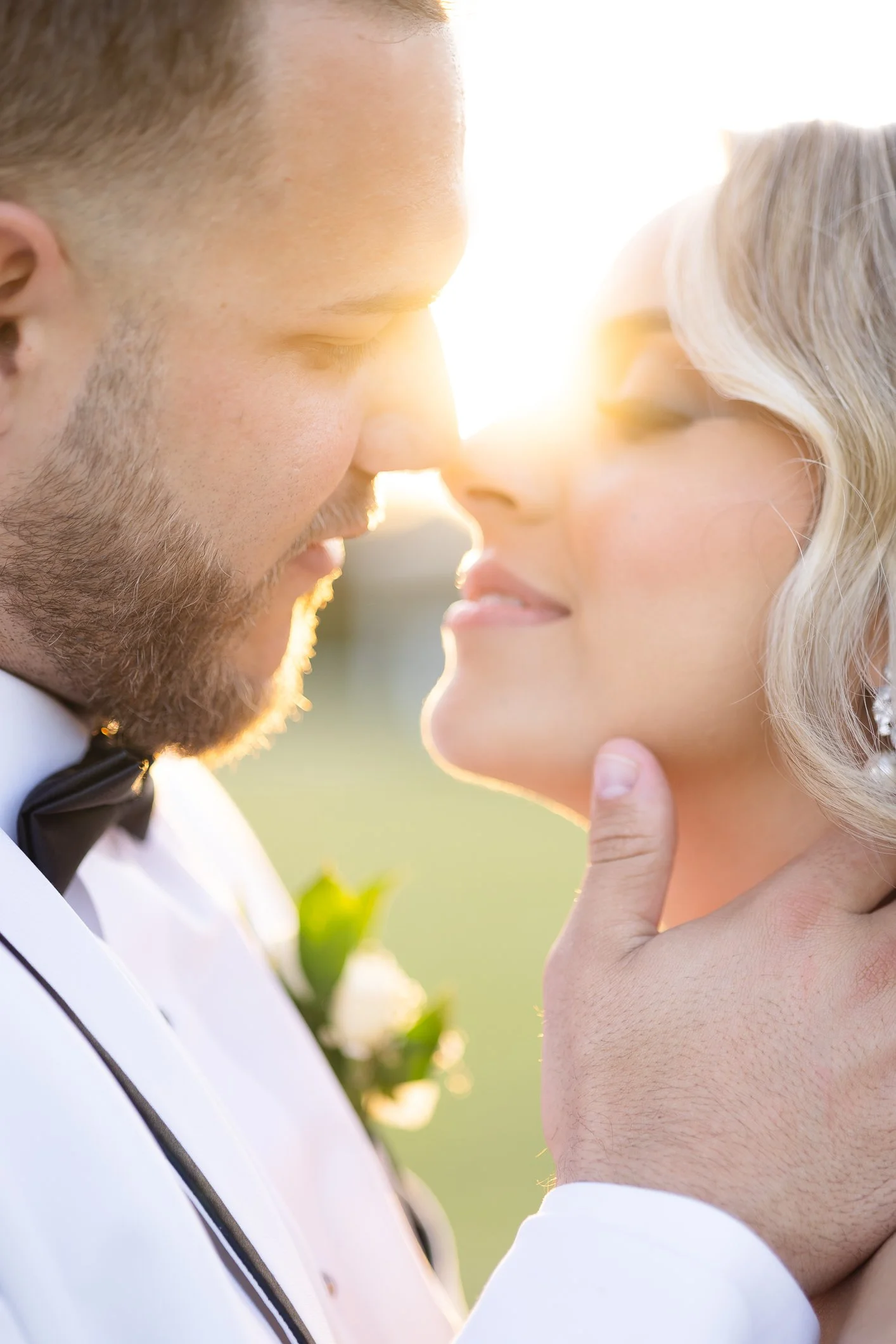 A couple in wedding attire with their foreheads touching, gazing at each other at sunset.