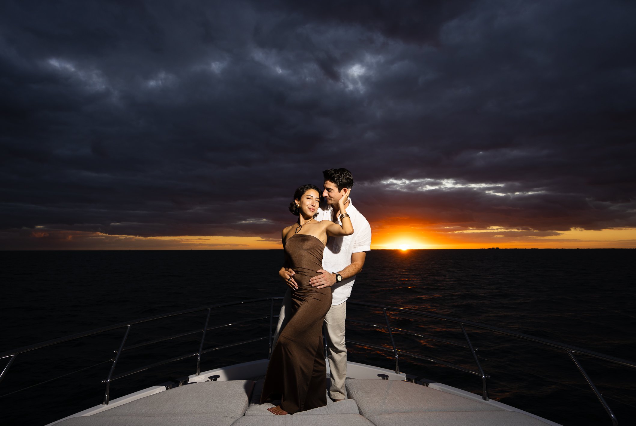 A couple stands on the bow of a boat at sunset, with dark clouds overhead and the ocean in the background.