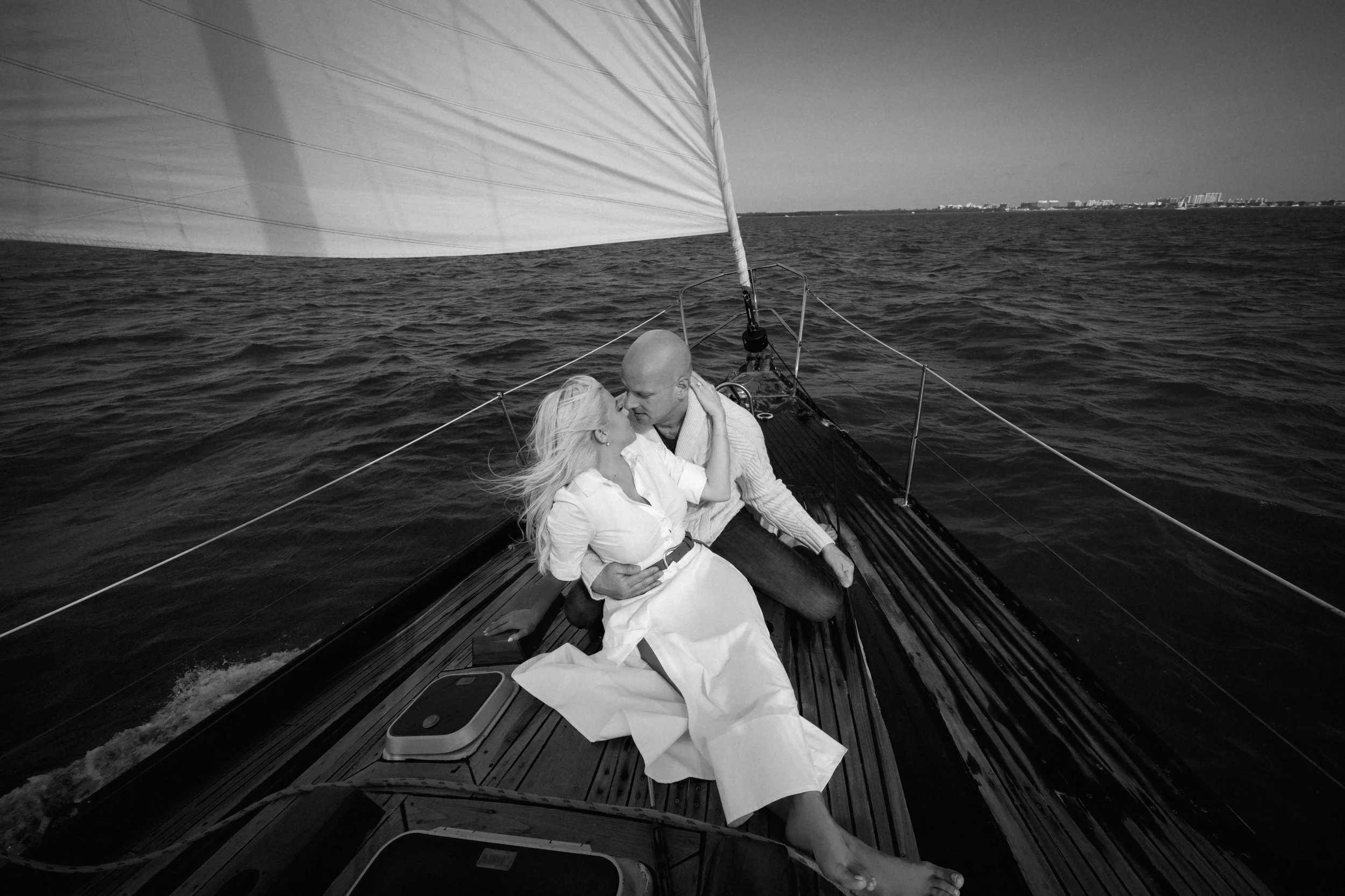 A black and white photo of a couple on a sailboat, sitting close and leaning in for a kiss, with the ocean and city skyline in the background.
