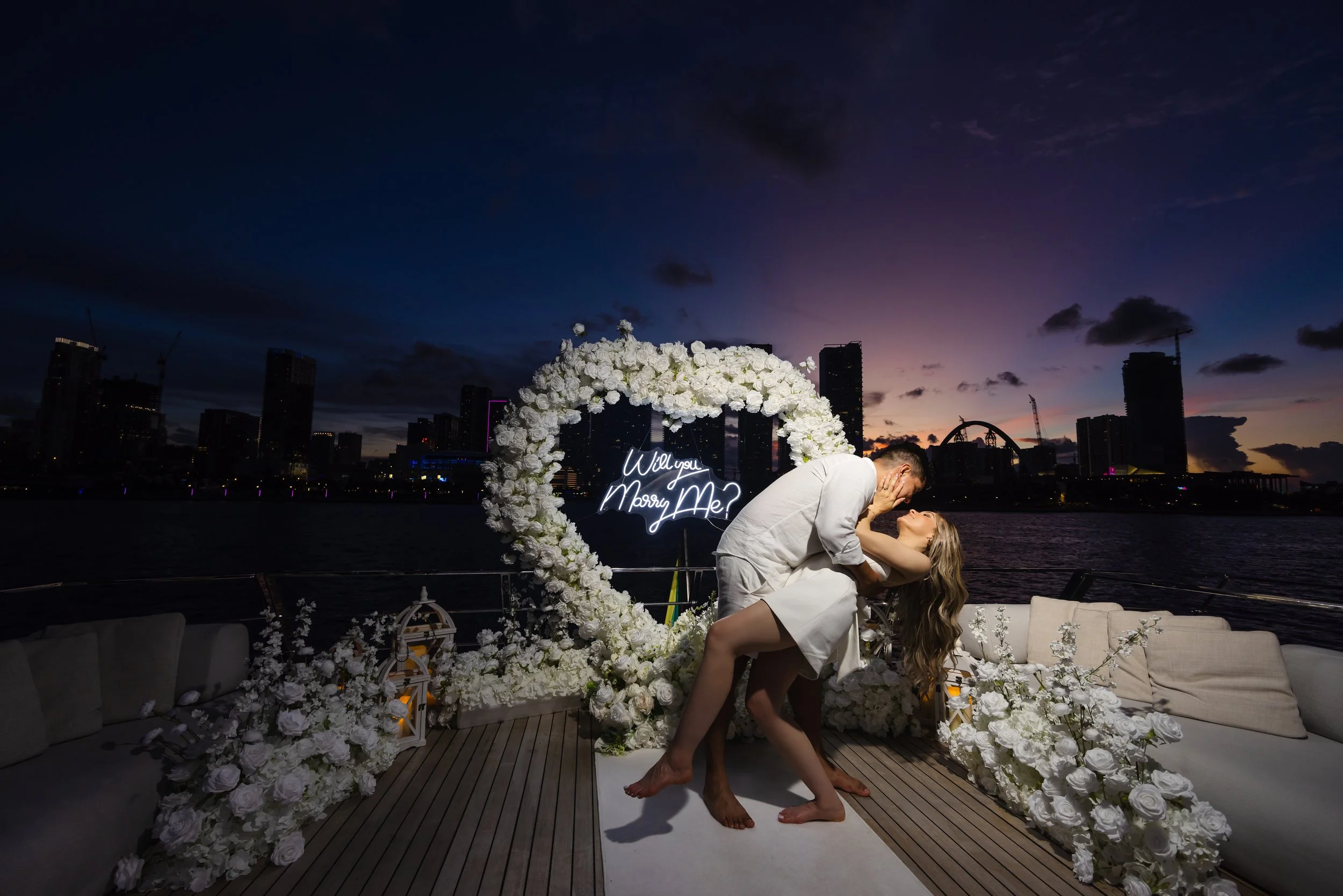 A couple kissing on a yacht during sunset with city skyline in the background, surrounded by white floral decorations and a neon sign that says, 'Will you marry me?'