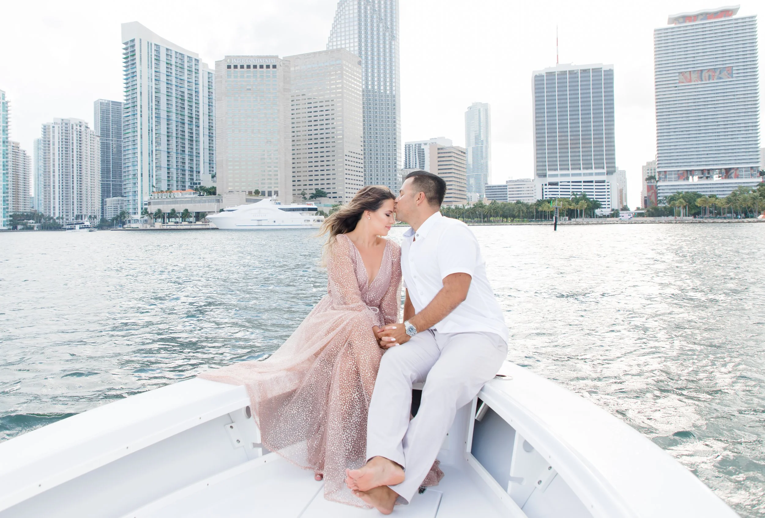 A couple sitting on the bow of a boat with a city skyline in the background, the man is kissing the woman's forehead, and they are holding hands.