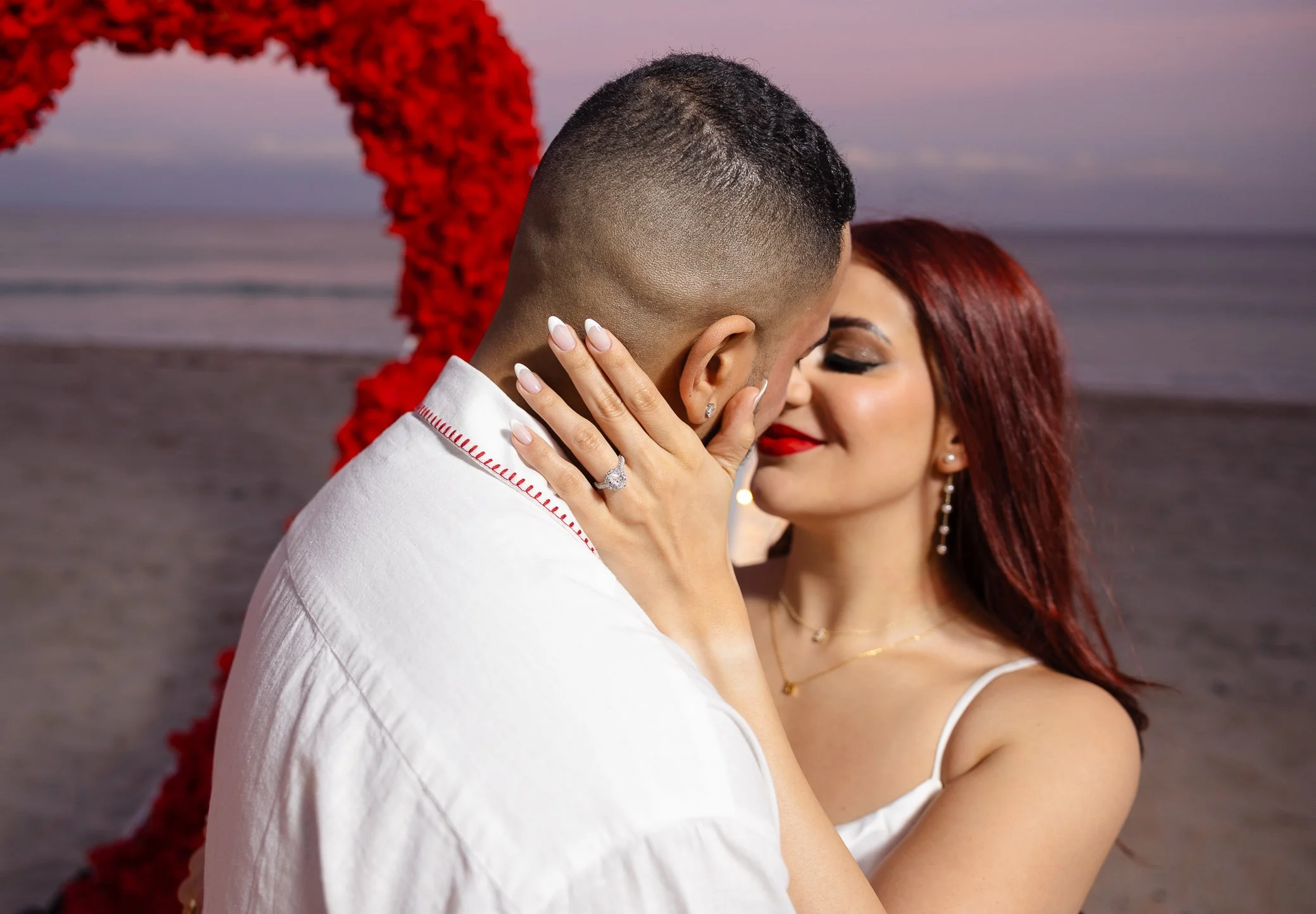 A couple sharing an intimate moment on the beach at sunset, with the woman holding the man's face and showing off an engagement ring.