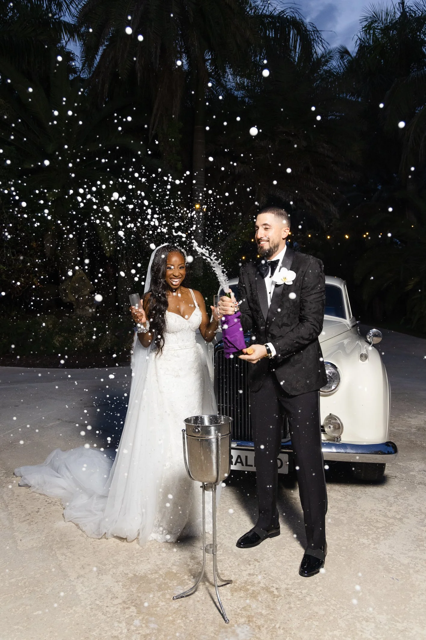 A bride and groom celebrate their wedding outdoors next to a vintage car, with champagne and confetti in the air.