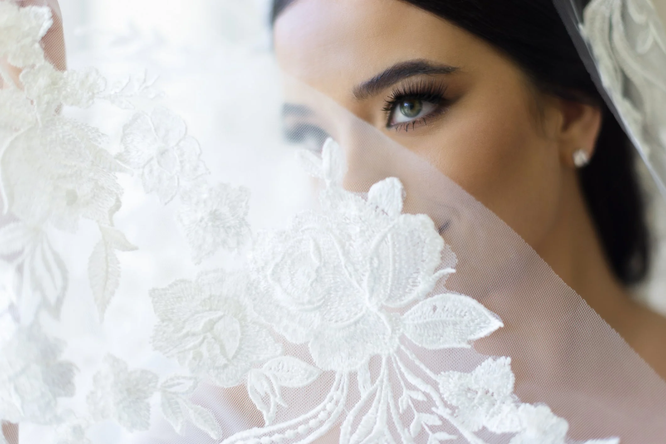 Close-up of a woman's face with striking eye makeup, looking through white lace floral fabric, possibly a bridal veil.