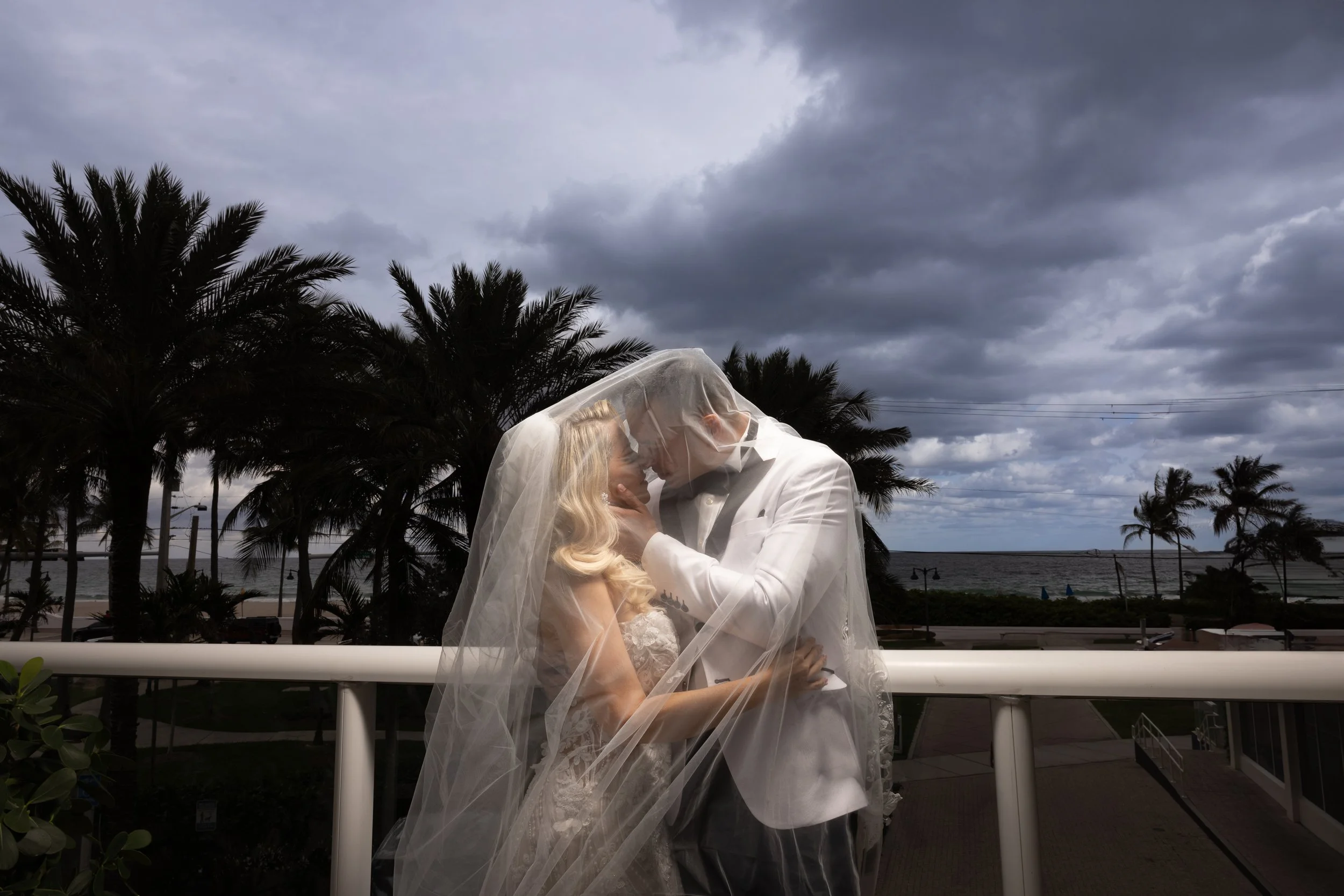 A bride and groom share a kiss under a veil during their wedding on a balcony overlooking palm trees, the ocean, and a cloudy sky.