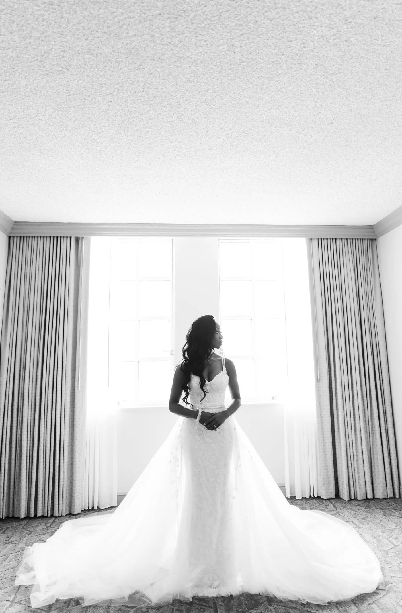 A woman in a wedding dress standing indoors near a large window with curtains.