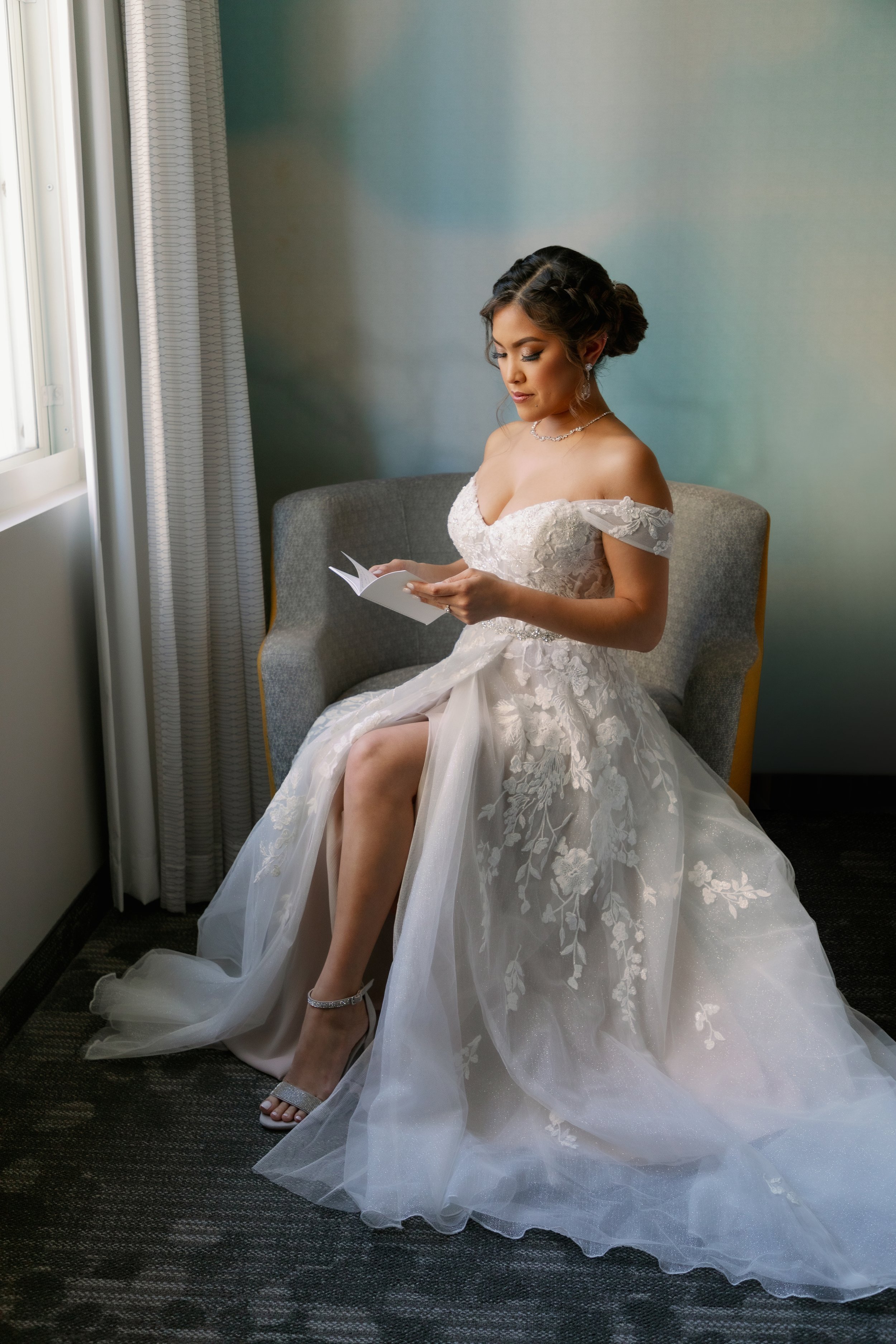 Bride sitting on a chair, reading a small book, in a wedding gown with lace details, near a window.