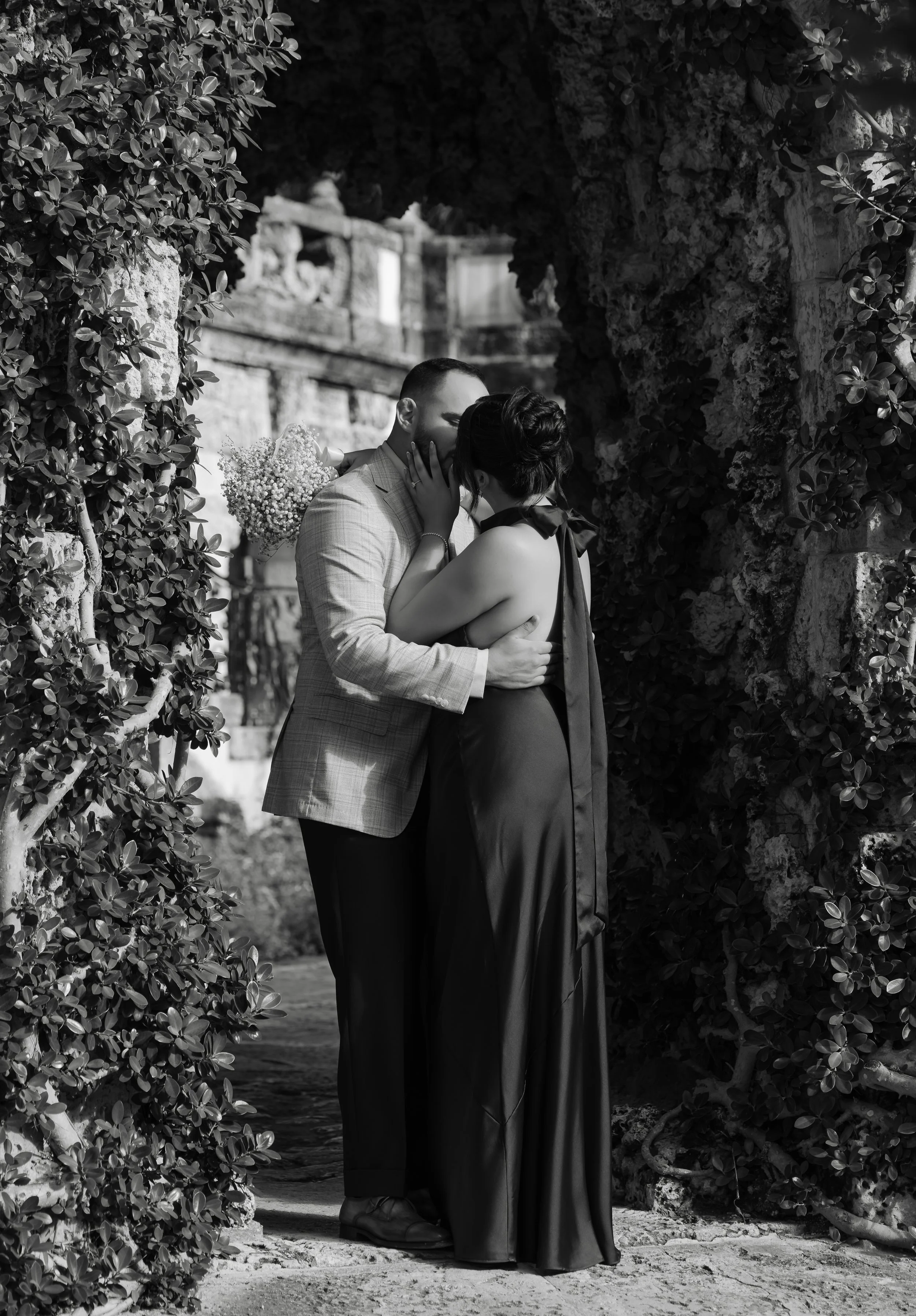 A couple sharing a romantic kiss in an outdoor setting framed by stone walls and foliage, with one holding a bouquet of flowers.